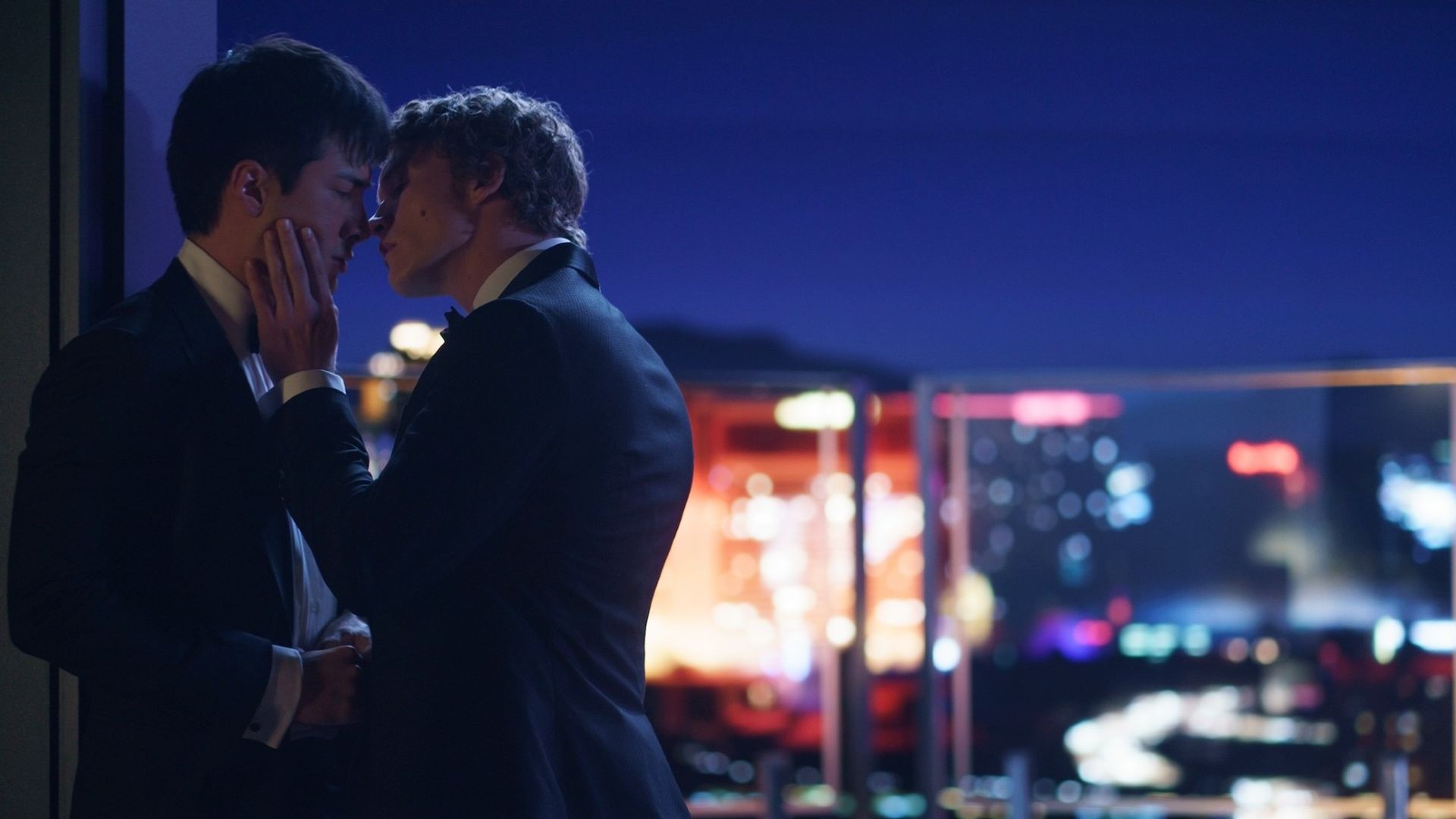 Two men in black tuxedos share an intimate moment on a balcony at night, city lights blurred in the background. One gently touches the other's face as they lean in.