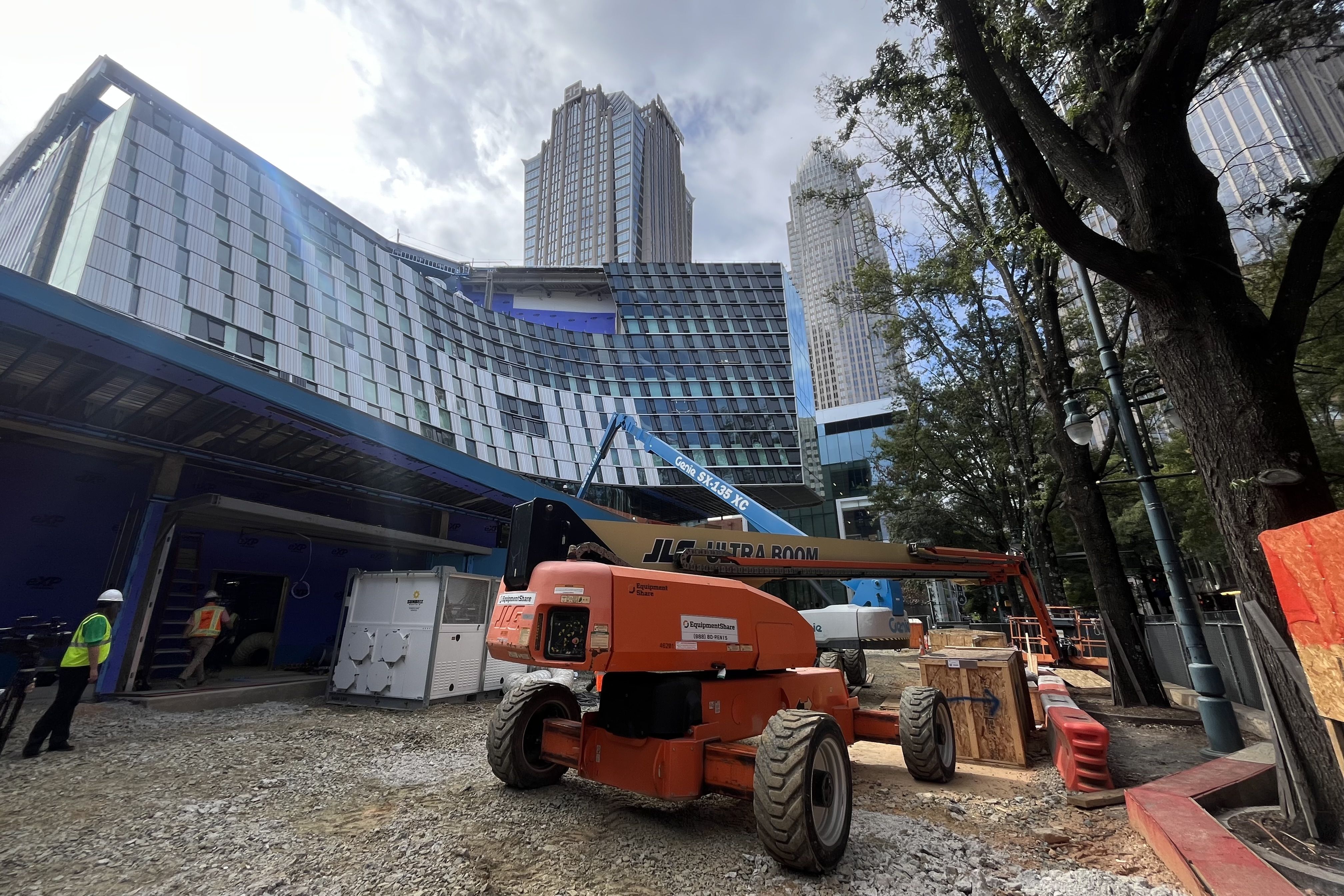 Construction site with a curved glass building under construction, orange and blue cranes, workers in safety vests, gravel ground, and tall skyscrapers and trees in the background.