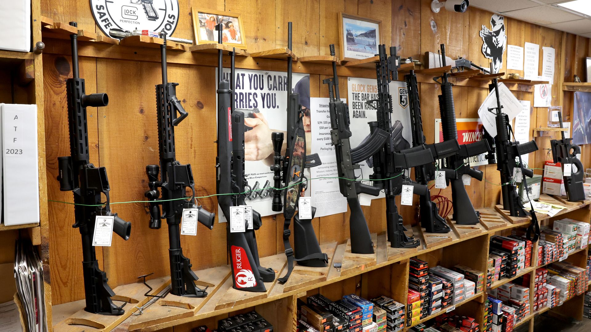 Indoor gun shop display: a wooden wall lined with multiple black rifles on racks, ammo boxes on lower shelves, posters and a large Glock sign above, and a security camera on the ceiling.