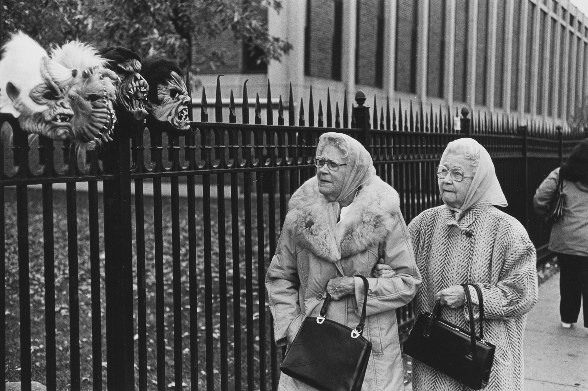 Black-and-white photo of two elderly women in coats and headscarves walking beside a tall wrought-iron fence, with grotesque monster masks mounted along the fence.