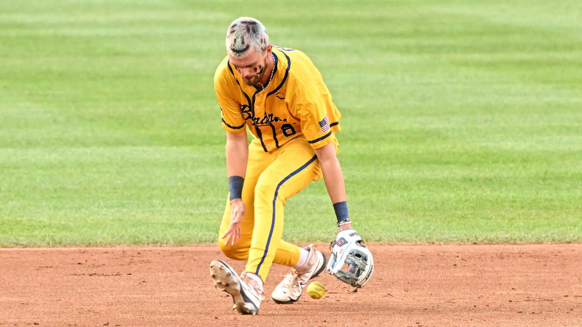Baseball player in a yellow uniform with black trim and an American flag patch fields a ground ball on a dirt infield with green grass in the background.