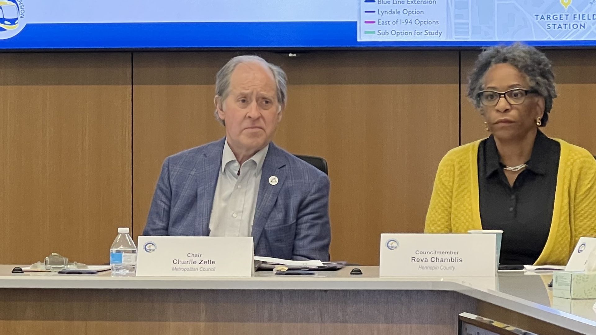 Meeting with officials seated at a conference table; a screen behind shows a map and text about West Broadway transit options, including routes and infrastructure challenges.