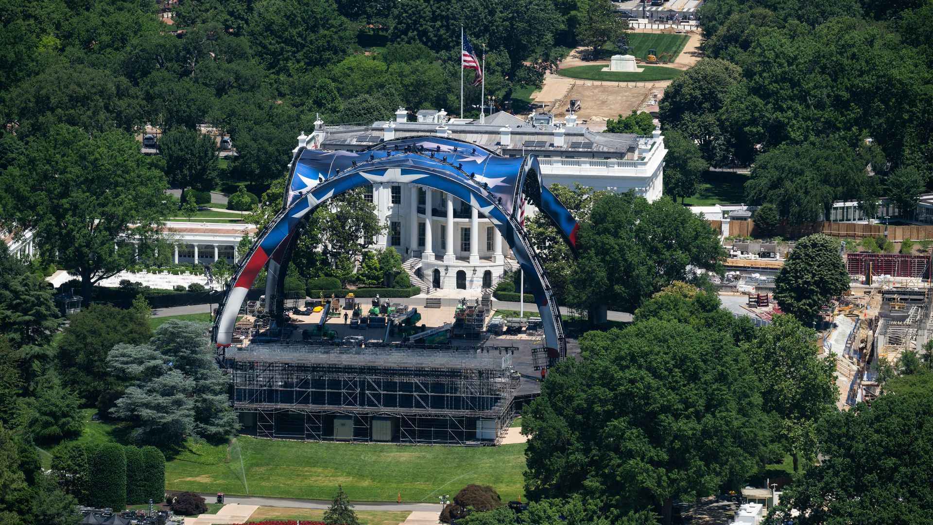 Bird's-eye view of a white neoclassical government building surrounded by trees, with a blue star-pattern arch stage in front, a tall American flag atop, and a circular fountain with red flowers in the foreground.