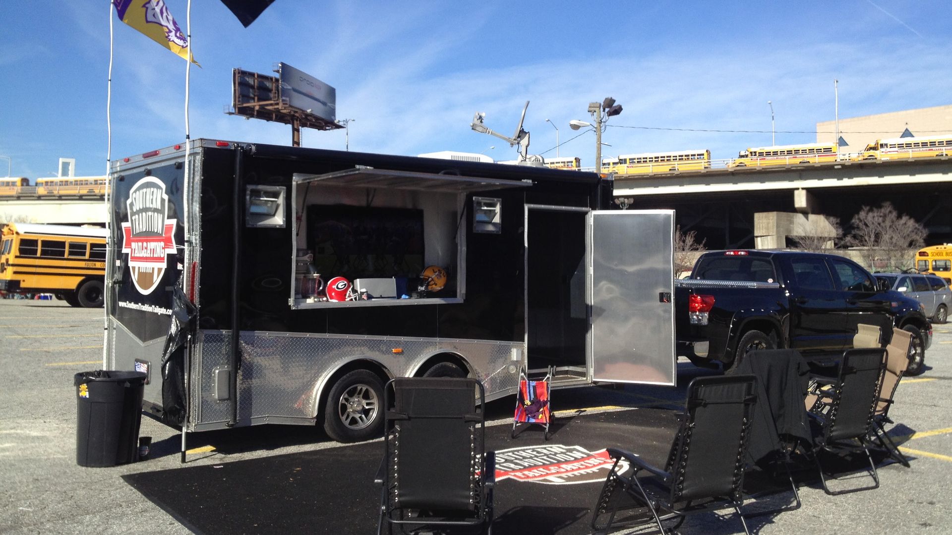 Black tailgating trailer with Southern Tradition Tailgating logo, two flags, and football helmets showcased, parked next to black pickup truck and folding chairs on a sunny day.
