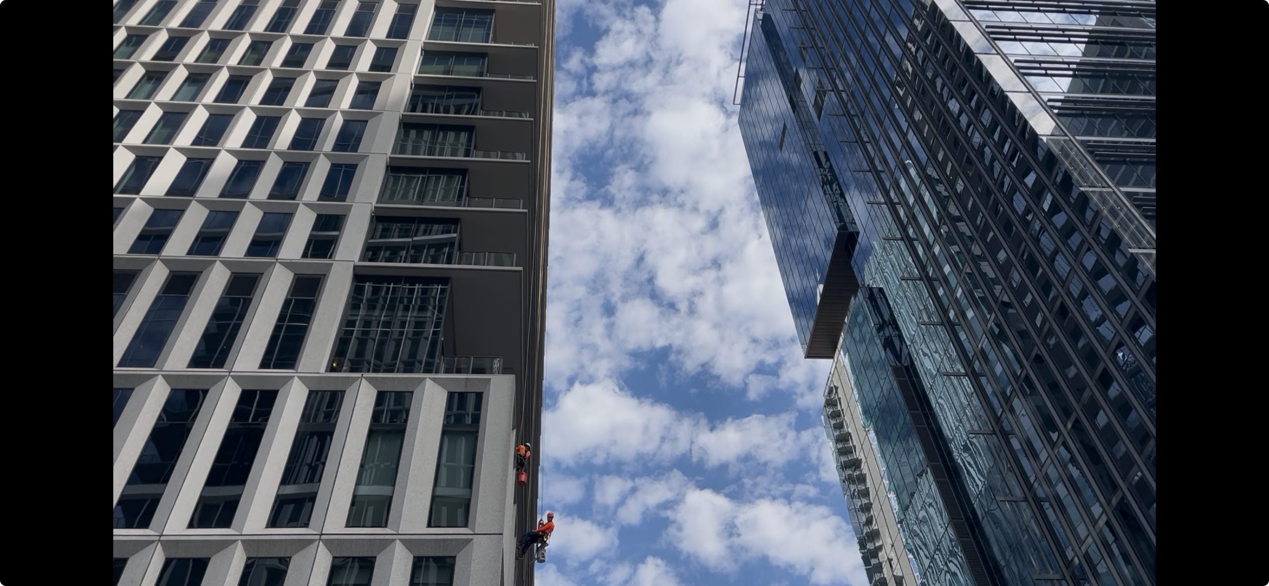 Two tall modern buildings frame a blue sky with clouds: left is white concrete with square windows, right is glass-faced skyscraper. A worker in orange safety gear hangs between them.