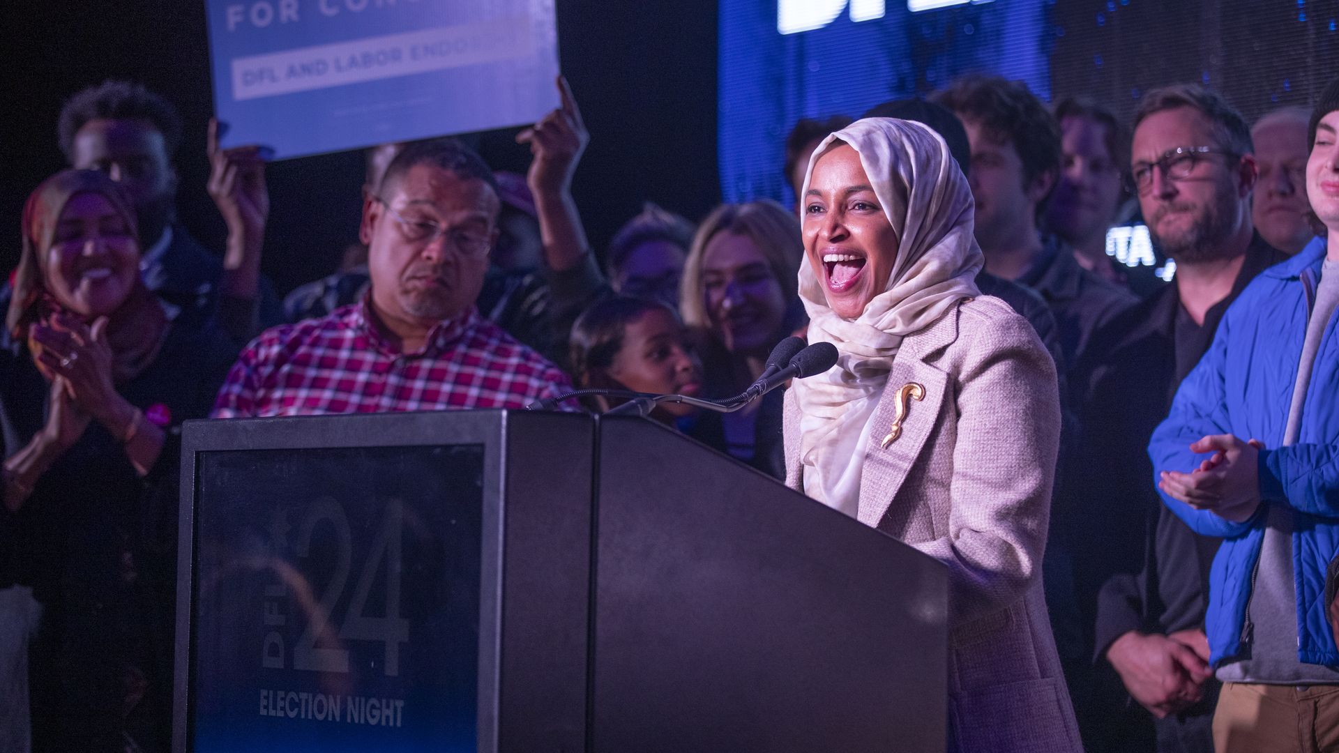 Ilhan Omar wearing a white hijab and suit while surrounded by a crowd.