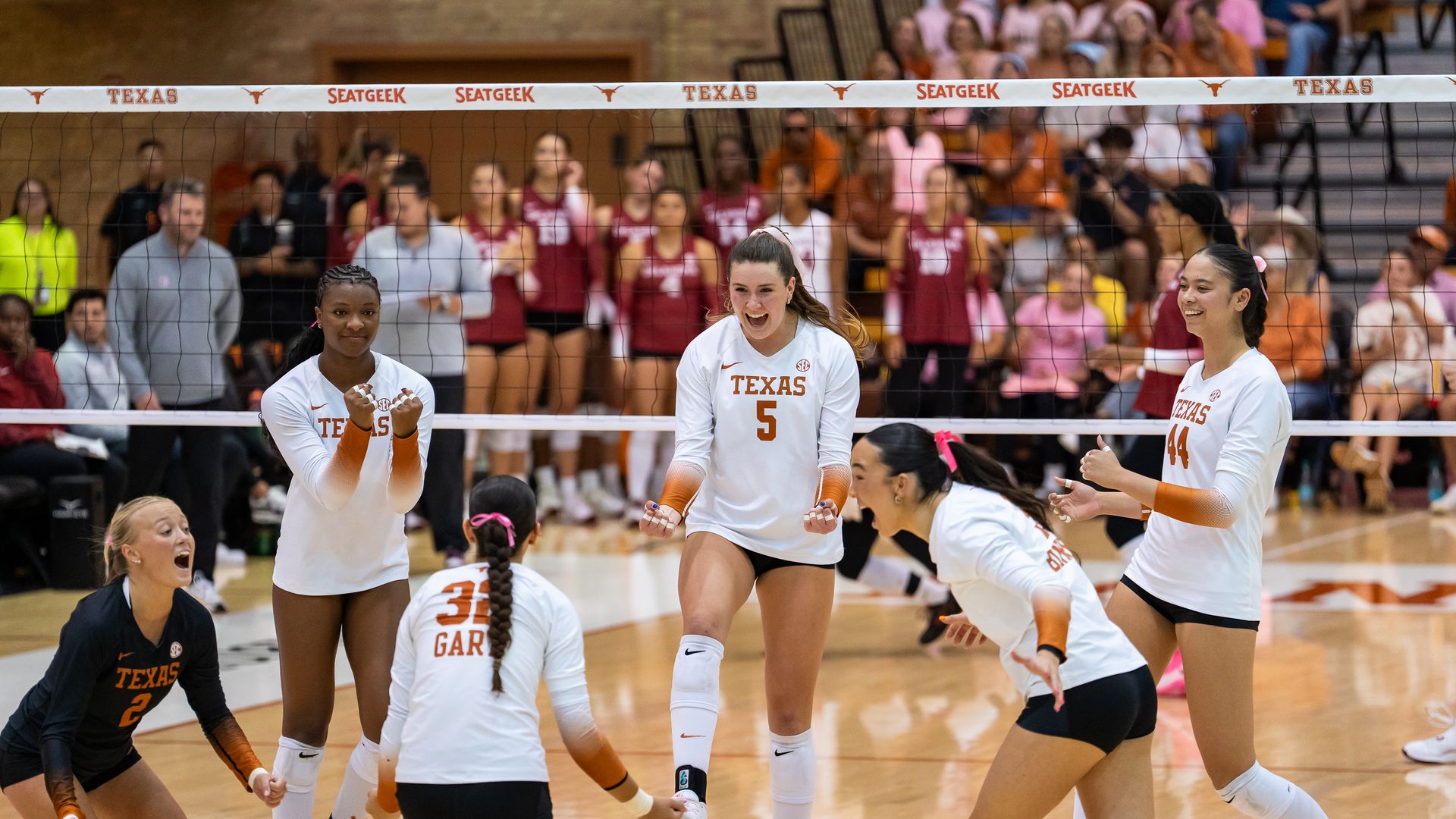 Texas women’s volleyball team in white uniforms celebrating on court with joyful expressions during a game, fans and opposing team in the background.