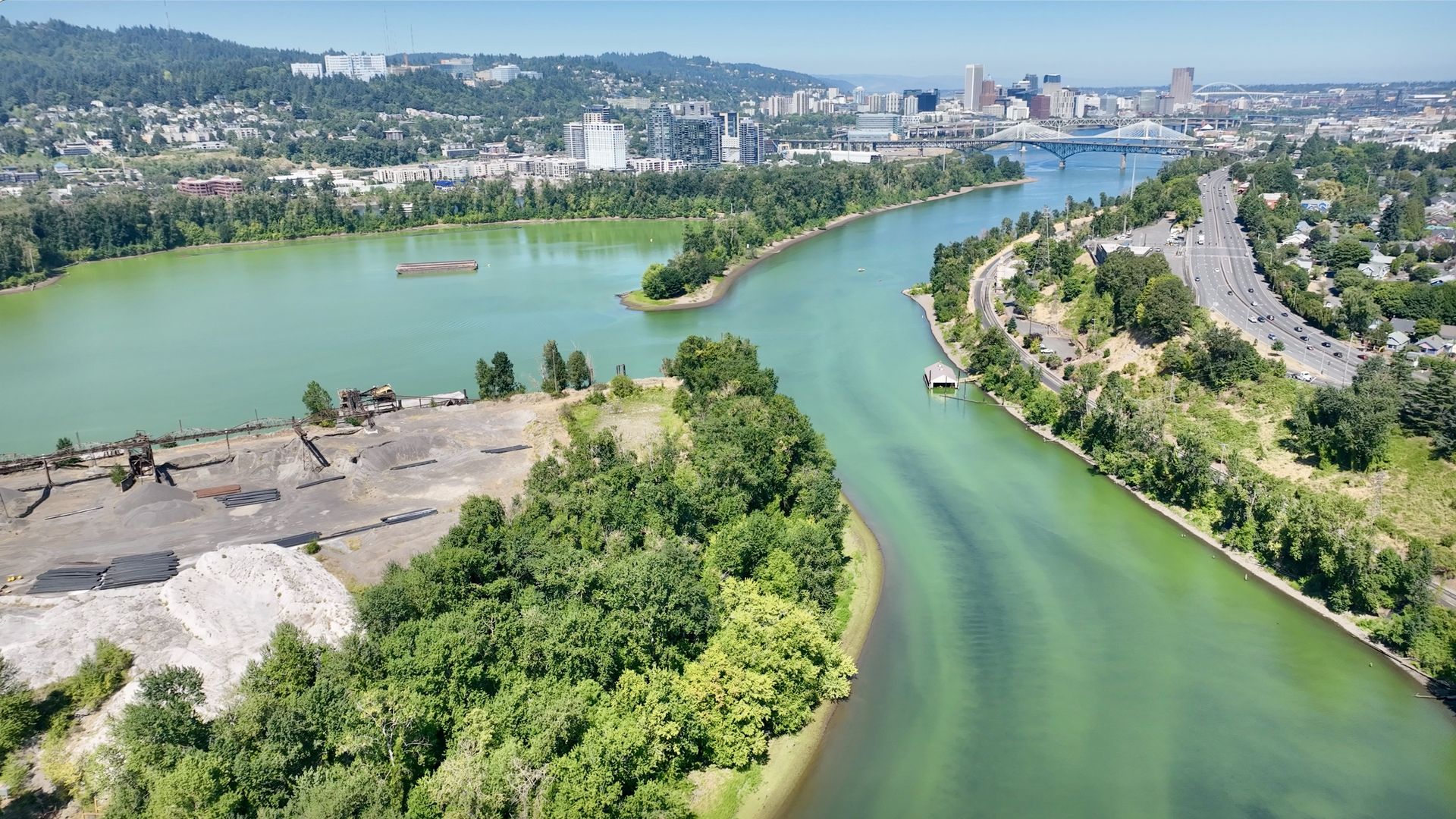 Aerial view of a green river curving through a city with a mix of dense trees, industrial area, highways, and urban buildings under a clear blue sky.