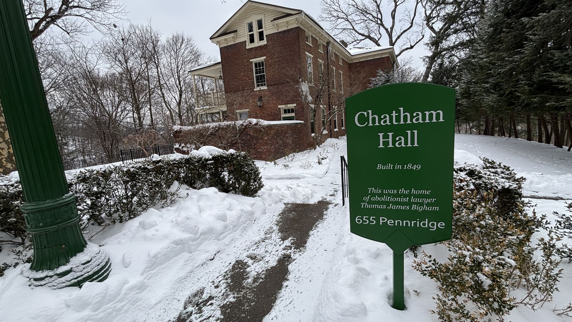 Snow-covered pathway and greenery lead to red brick building under gray sky. Green sign reads "Chatham Hall, Built in 1849, home of abolitionist lawyer Thomas James Bigham, 655 Pennridge."