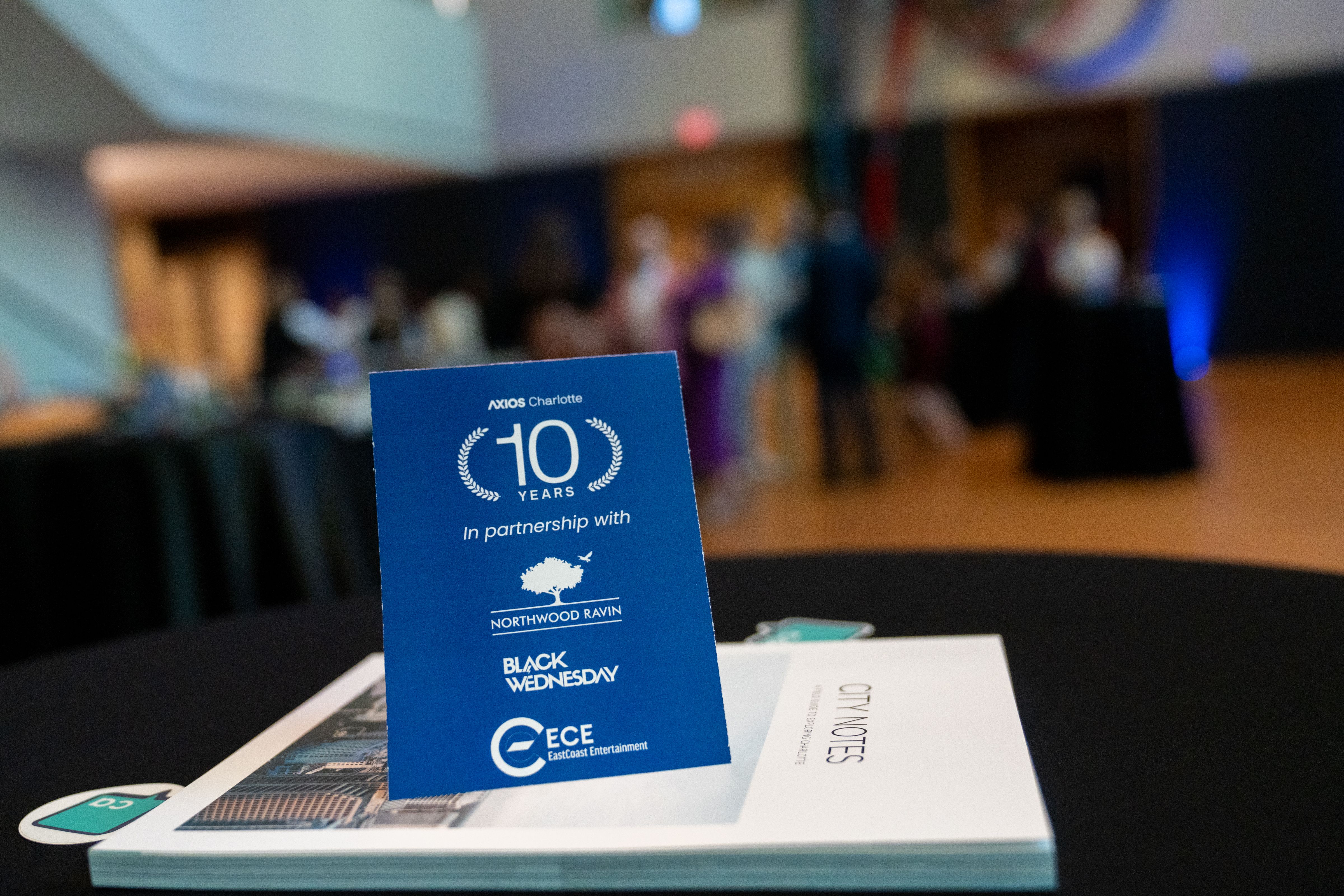 Blue event card on a table reads "AXIOS Charlotte 10 years in partnership with Northwood Ravin, Black Wednesday, East Coast Entertainment." Blurred background with people and dim lighting.