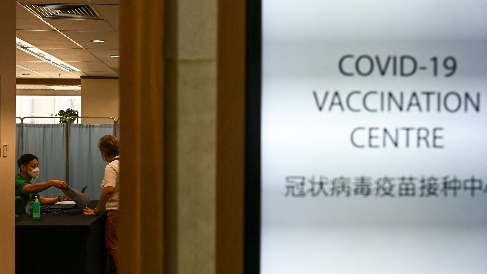 Photo of two people speaking in a room while a sign on the outside says "COVID-19 Vaccination Centre" in English and Chinese