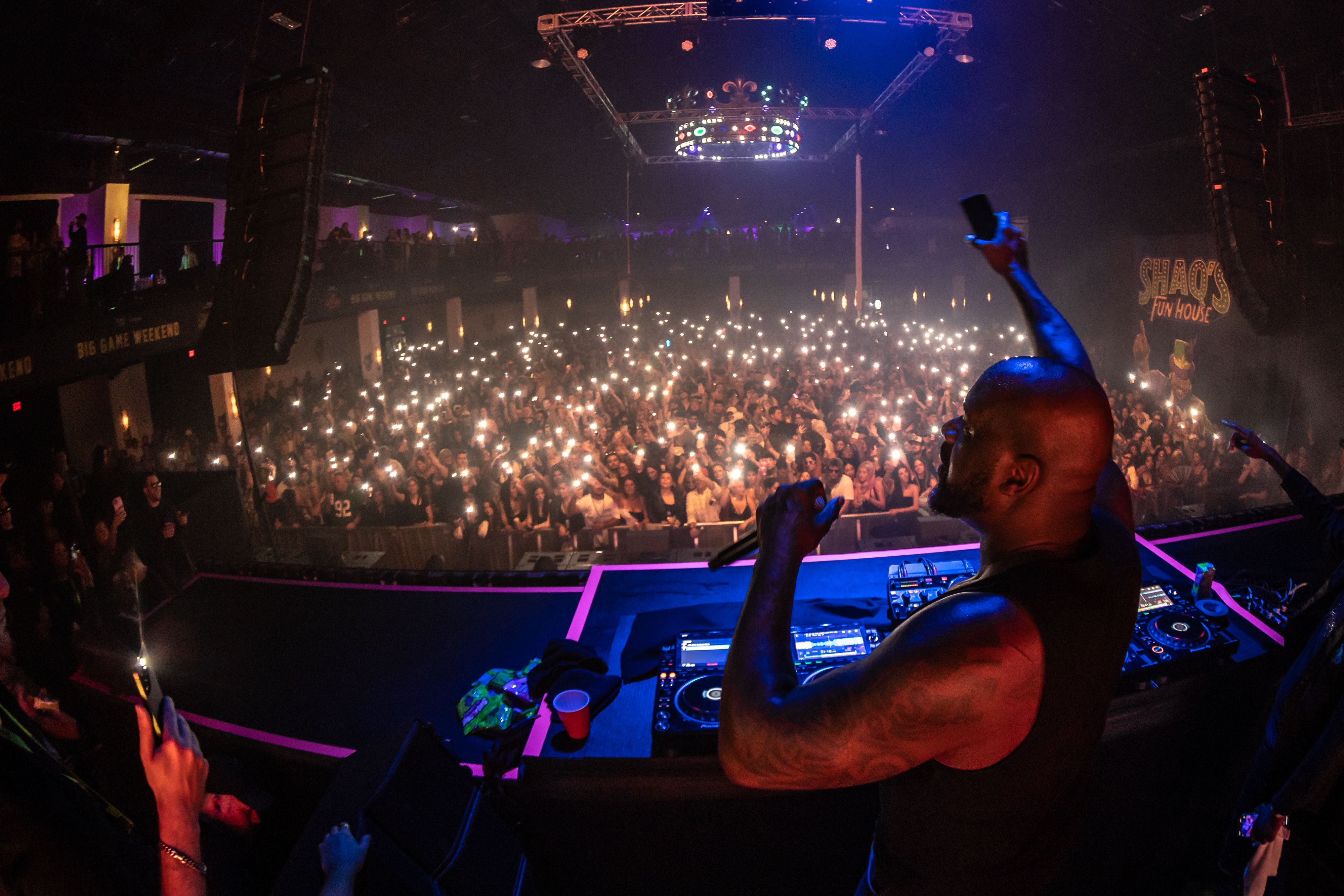 Shaq is viewed DJing from behind, with one hand in the air in front of a crowded audience.