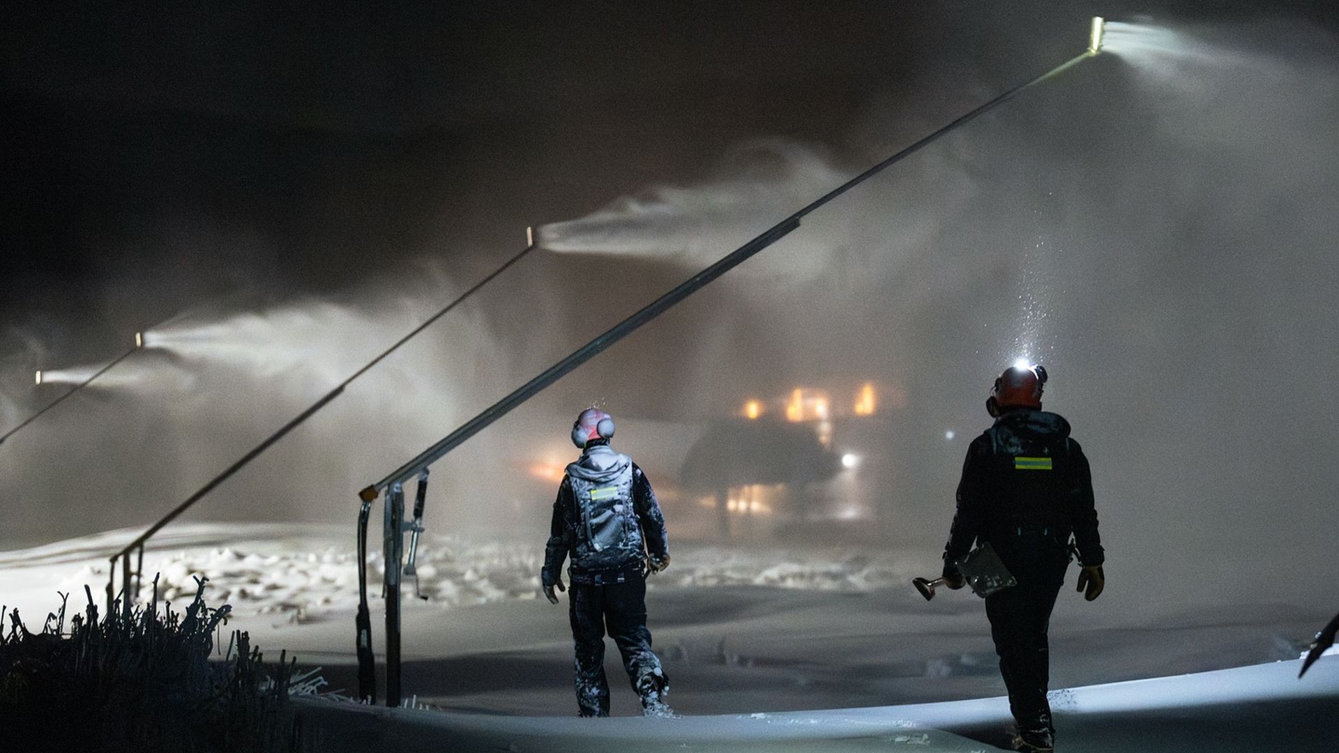 Two people in snow gear walking at night near machines spraying snow under dim lighting, with visible footprints in the snow and mist in the air.