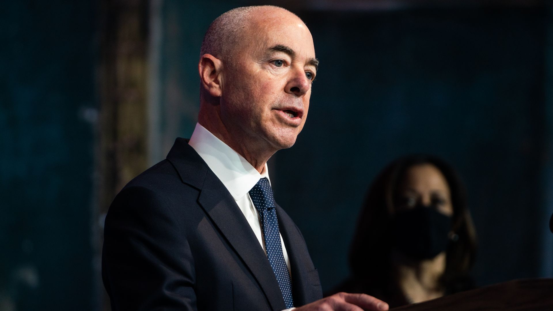Alejandro Mayorkas in a suite and tie speaking at an event with VP-elect Kamala Harris masked and standing in the background. 