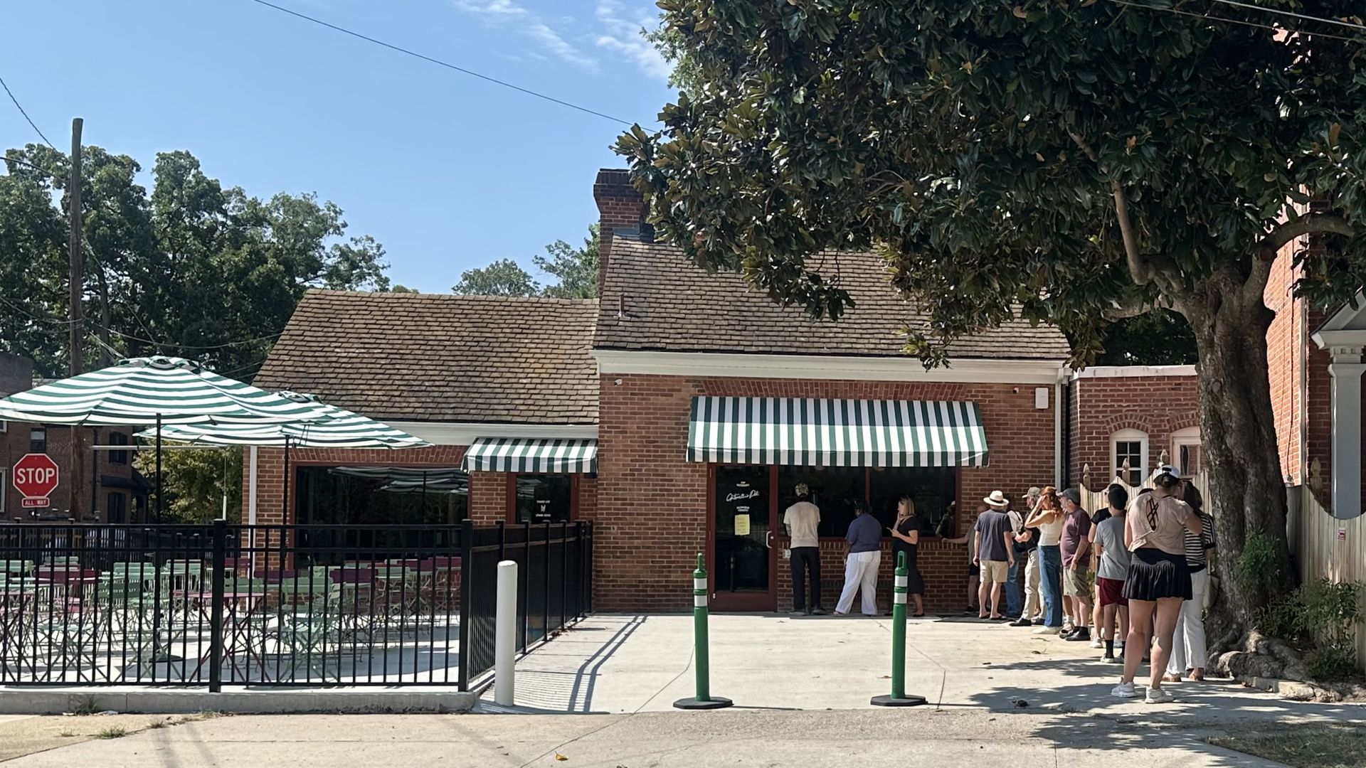 Sunny street scene with a brick storefront, green-and-white striped awnings, and outdoor seating under a striped canopy. People line up by the door as a large tree fills the right side; stop sign on the left.