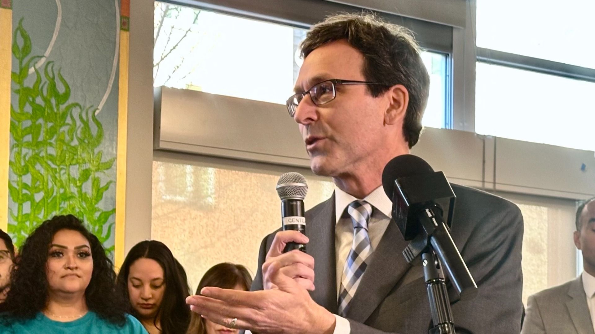 A man in a gray suit and striped tie speaks into a handheld microphone at a public event, with a crowd behind him and a green plant mural near glass windows and a second mic on a stand.