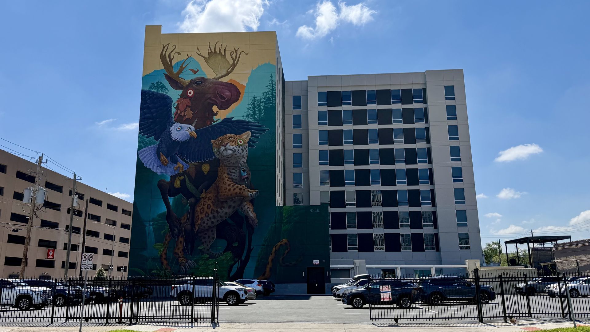 Blue sky over a tall mural-covered building showing a moose with antlers, an eagle, and a leopard, with mountains in the background. A modern glass building sits to the right; a parking lot ahead.