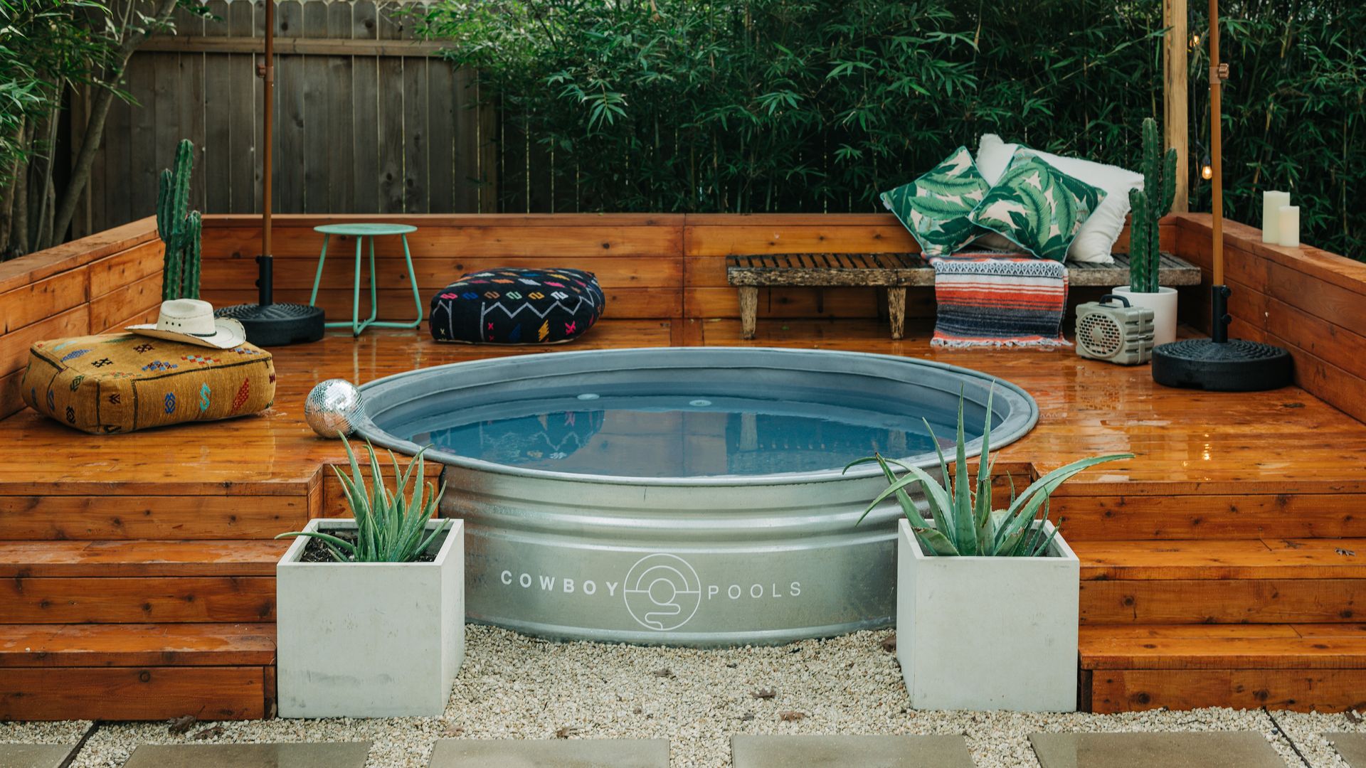 A circular metal pool with a Cowboy Pools logo on it filled with water surrounded by a wooden deck flanked by two square-shaped pots with spiky succulents inside. On the porch is a bench, table, cushion with a cowboy hat on top, cactus and two umbrellas. Behind it is a wooden fence and bushes.