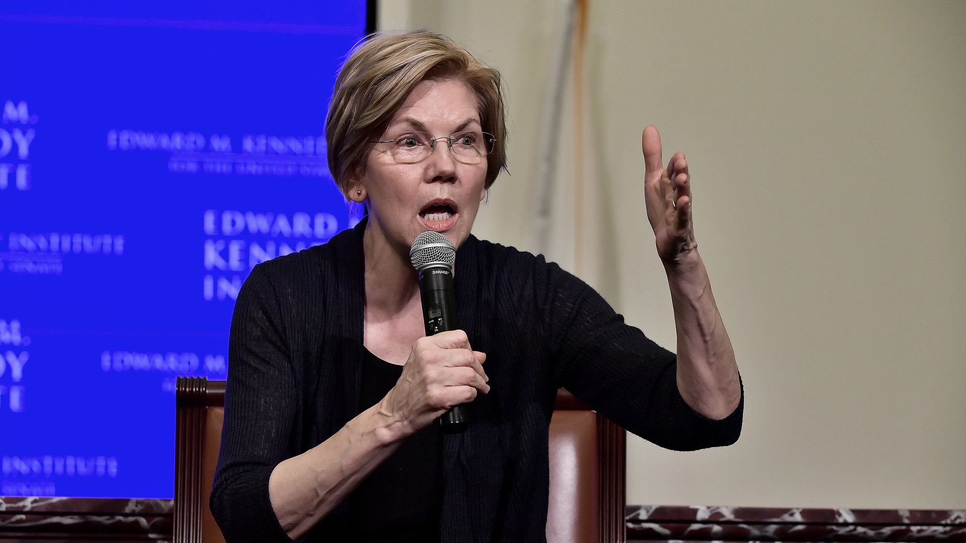 Senator Liz Warren gestures while holding a microphone, wearing black, before a blue and white background.