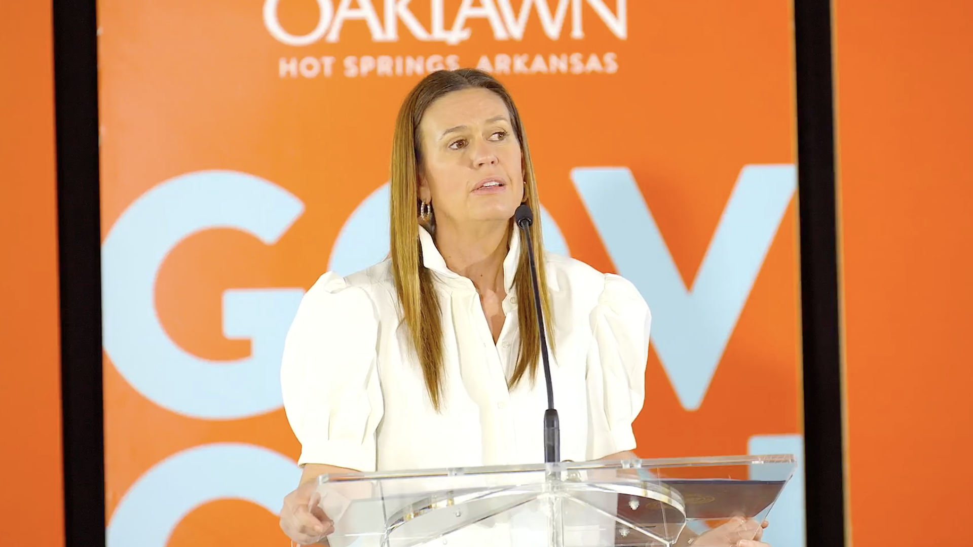 A photo of Republican Gov. Sarah Huckabee Sanders standing at a clear podium, wearing a white blouse and standing in front of an orange backdrop.
