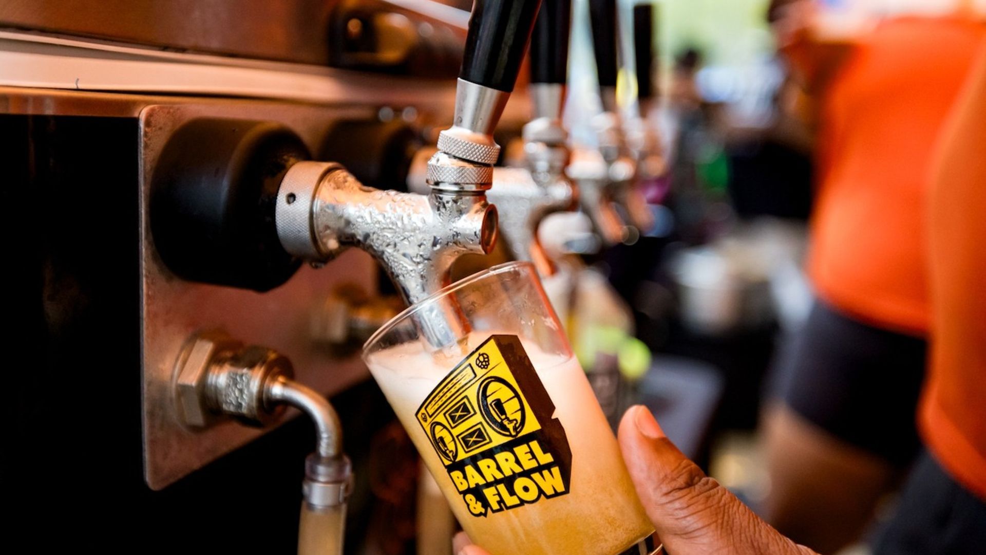 Close-up of a hand filling a glass with beer from a shiny metal tap. The glass has a yellow and black logo reading "BARREL & FLOW." The background is blurred with warm tones.