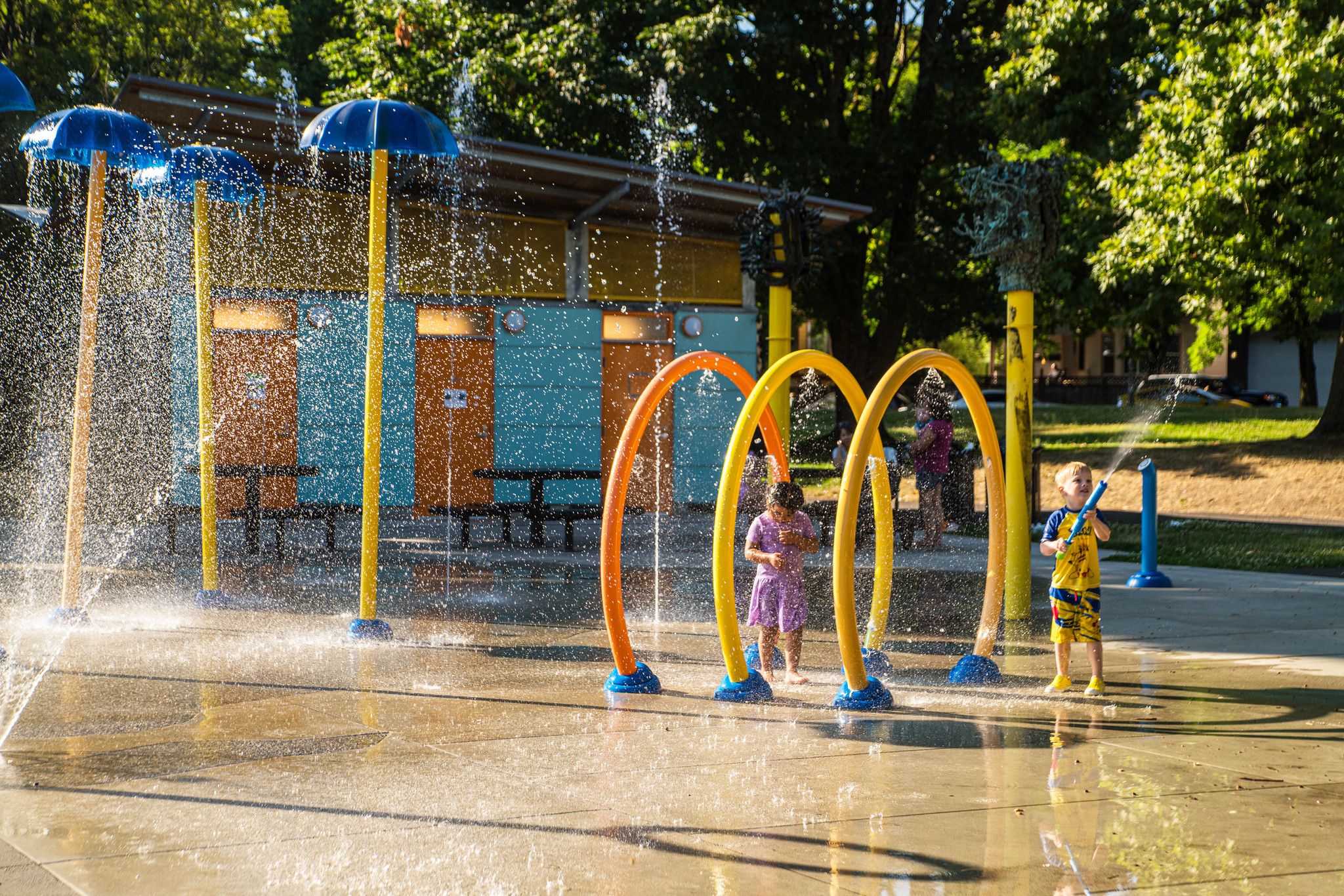 Two children play in a tunnel of sculptural elements that are yellow and orange in the Pratt Park spray park, while water sprays around them.
