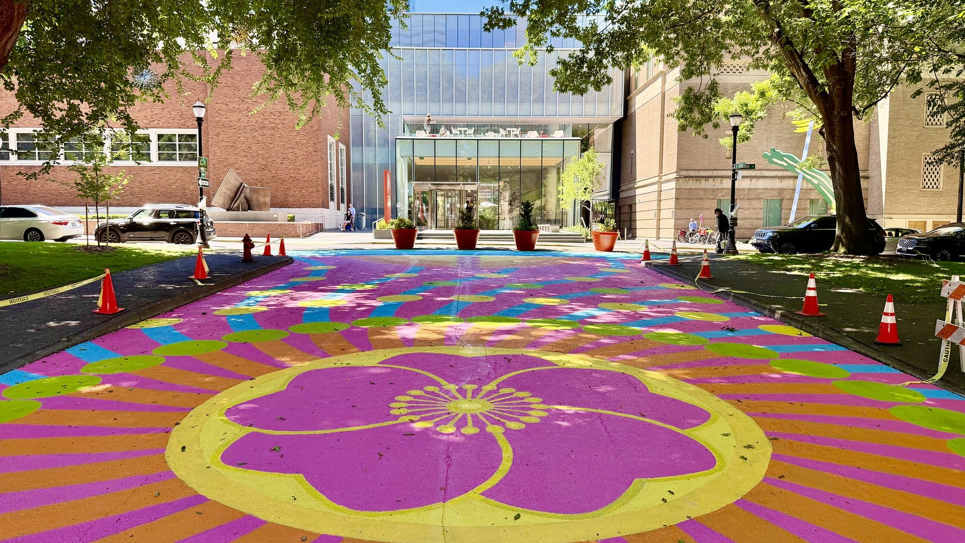 Modern glass-faced building between green trees. A vibrant rainbow circular mural covers the street with pink, purple, yellow, and green, bordered by orange cones; a few people near the entrance.