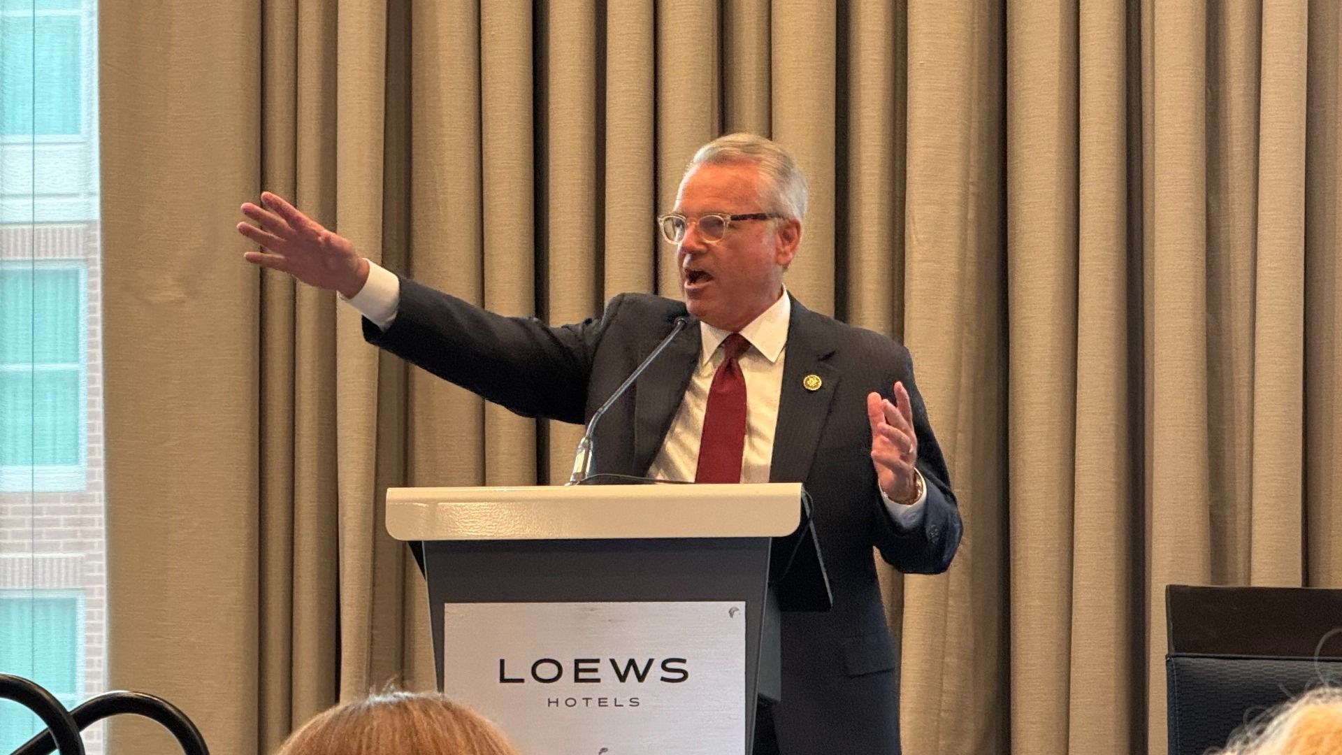 A man in a dark suit and red tie speaks at a LOEWS Hotels podium, gesturing with his left arm extended; microphone and beige curtains behind him.
