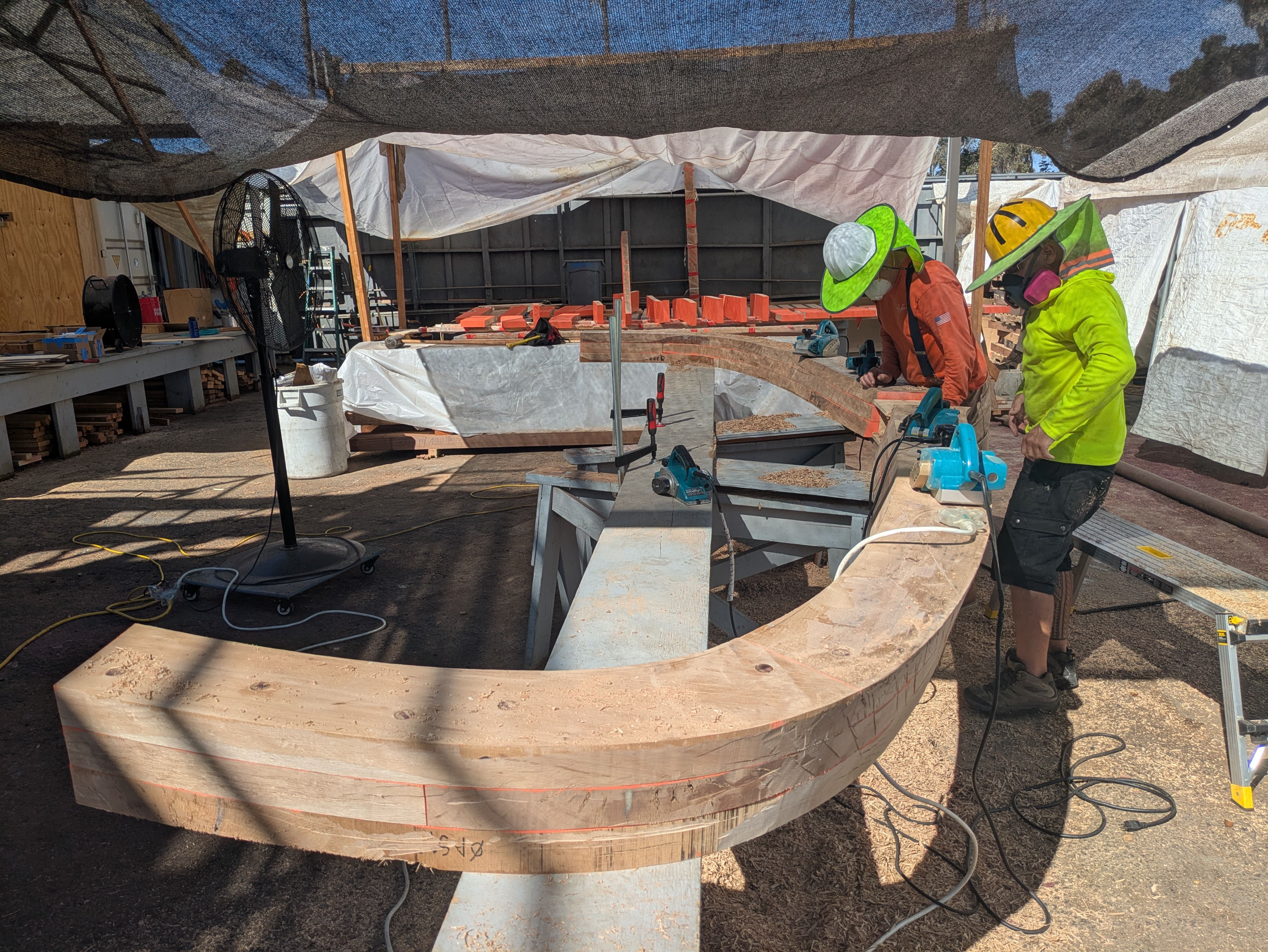 Two workers in neon safety gear and hard hats work at a ring-shaped curved wooden formwork under a shaded tarp, with orange bricks, tools, and a large fan nearby on a construction site.