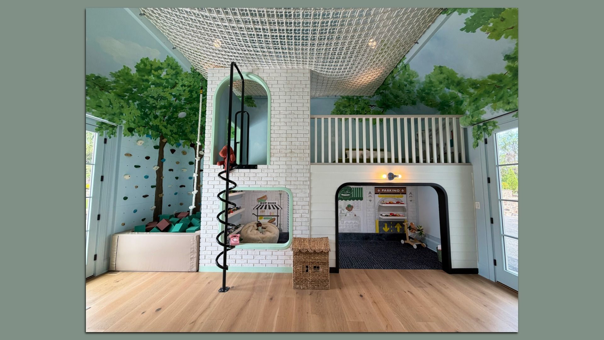 Indoor playroom with a white brick pillar, arched window inset, black spiral pole, climbing wall with colorful holds, tree mural, netted ceiling, and a loft with white railing above on a wooden floor.