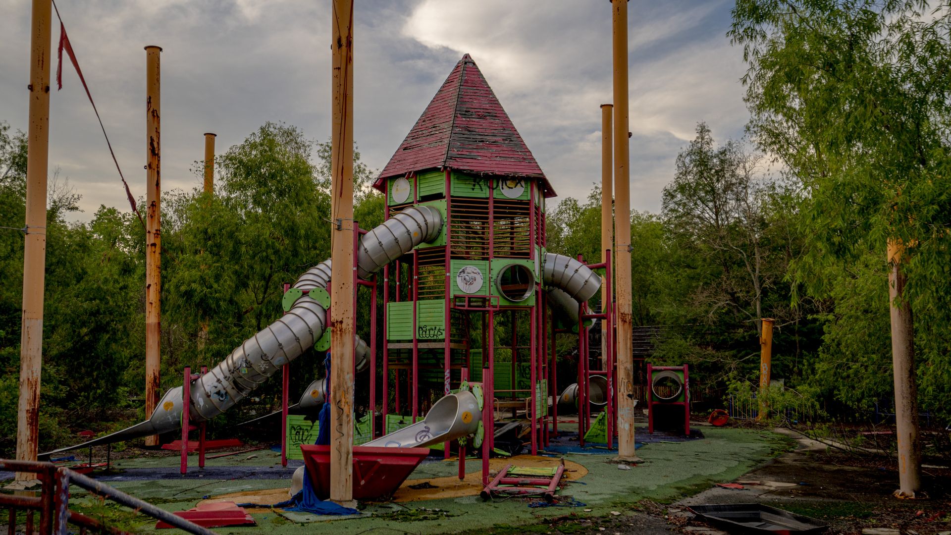Photo shows an abandoned slide playground with trees in the background.