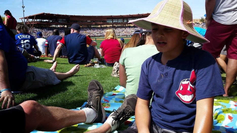 A young boy sits in the sun on the grass outside of a baseball field.