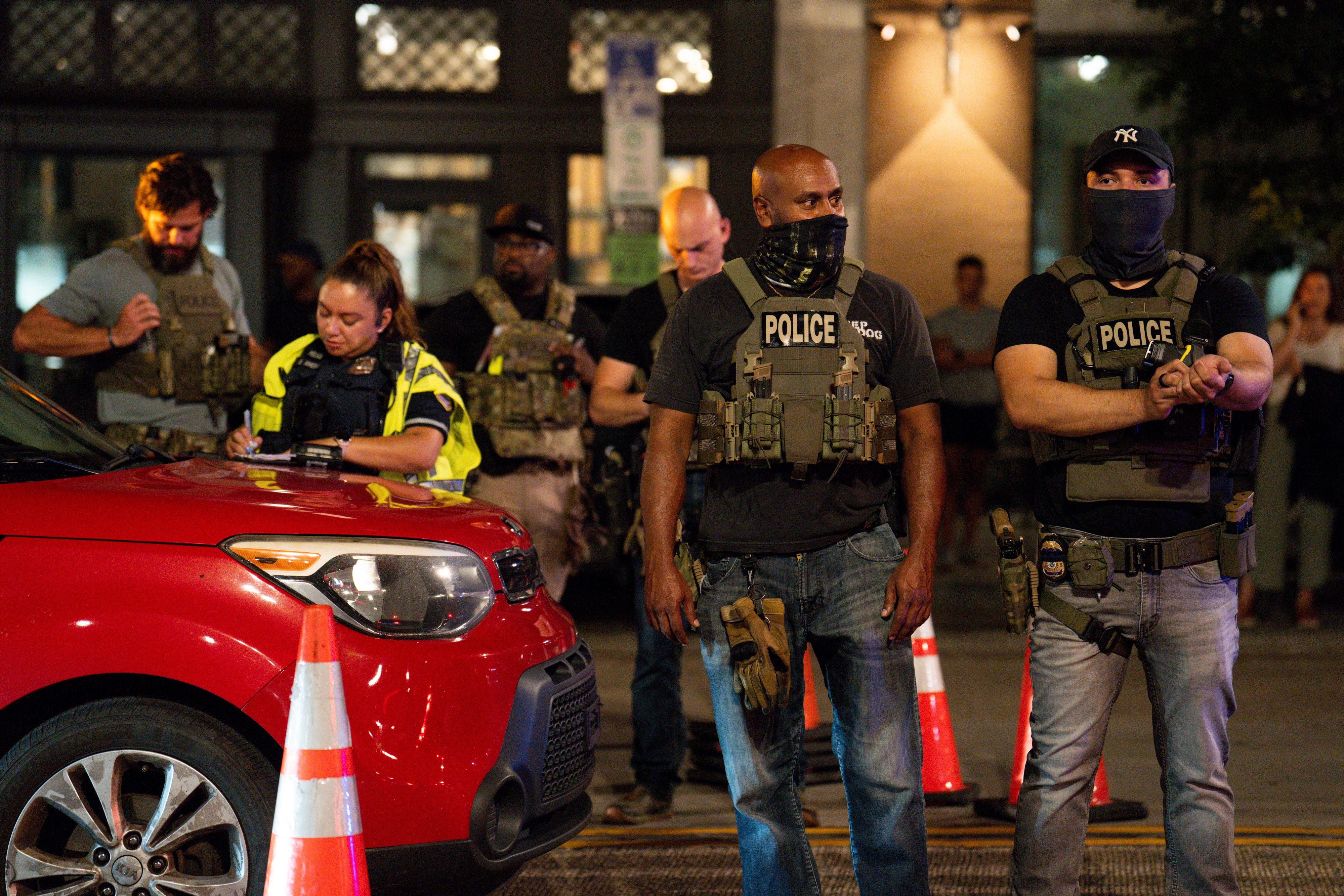 Police officers in tactical gear standing and working near a red car and orange traffic cones at night, with some officers wearing masks and vests labeled "POLICE".