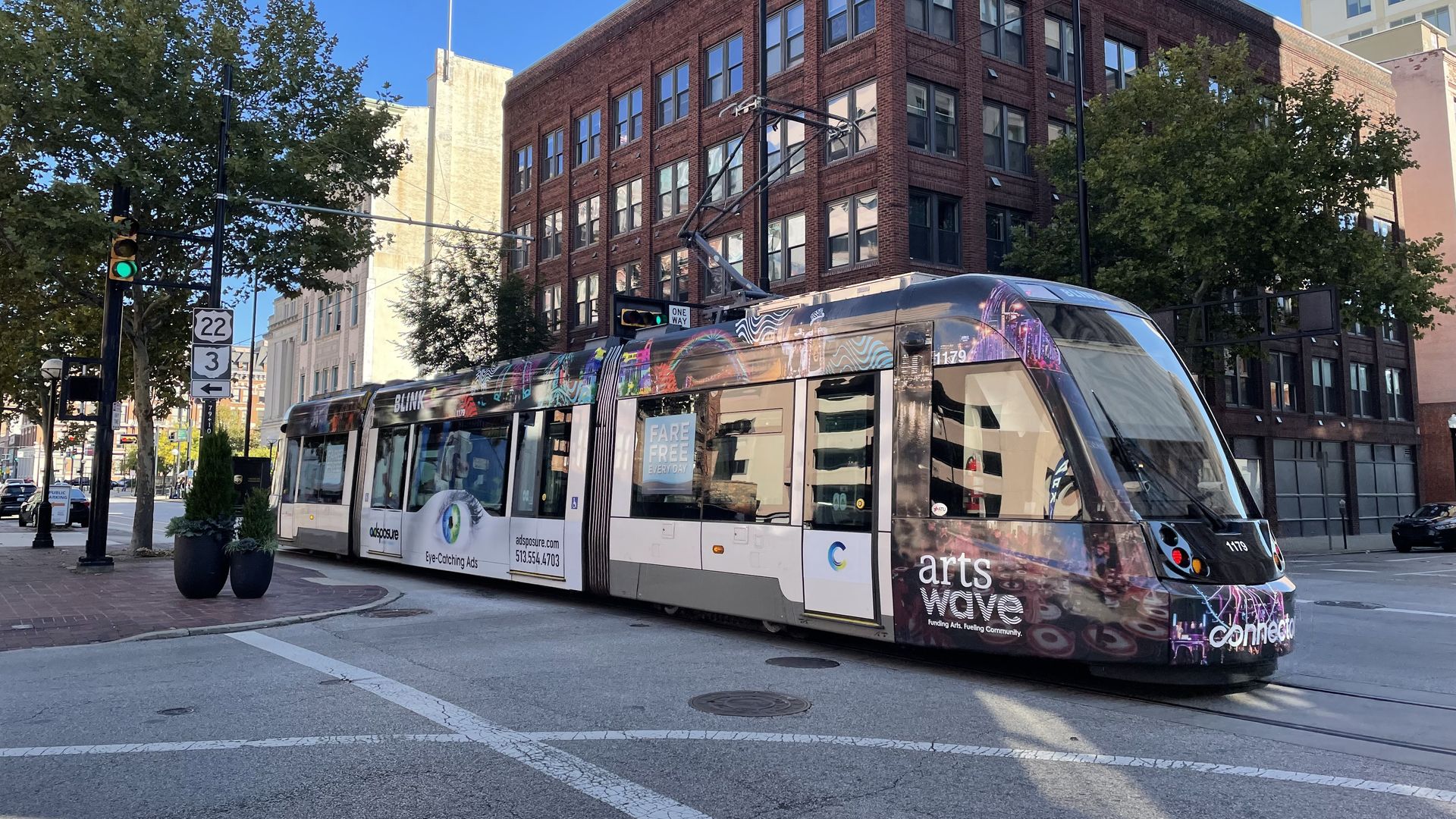 Urban street scene with a colorful tram in front of a brick four-story corner building, clear blue sky, and trees along the sidewalk; visible traffic signals and street signs.