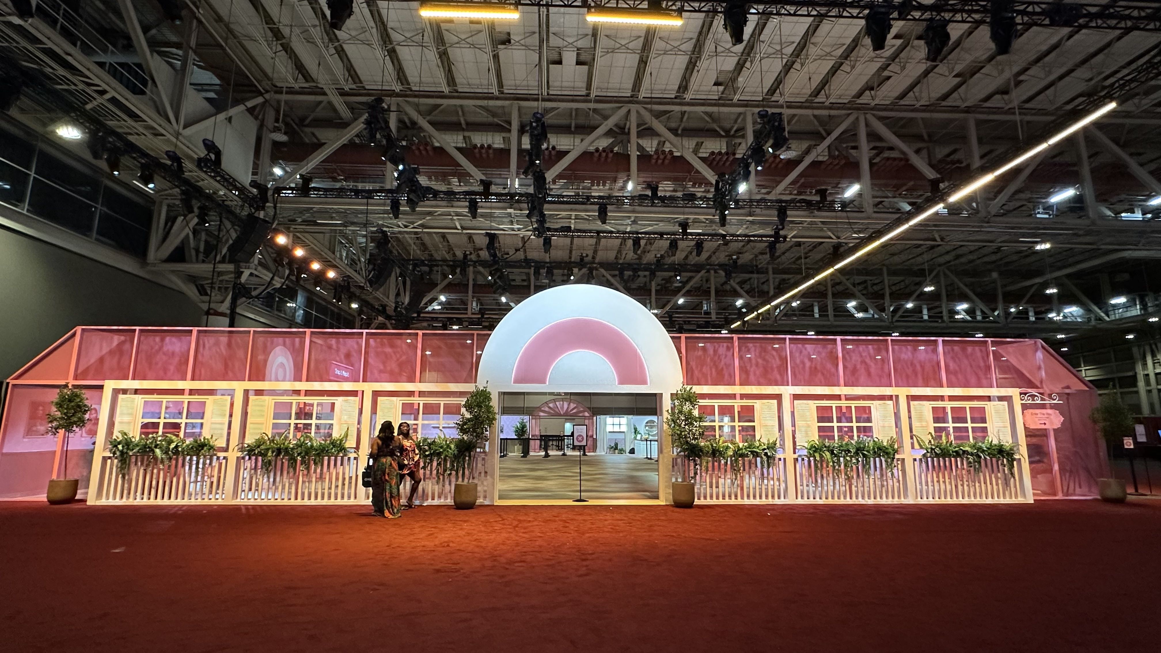 Large indoor pink and white structure resembling a house facade with plants, picket fence, and a large rainbow-shaped arch entrance under industrial ceiling lights.