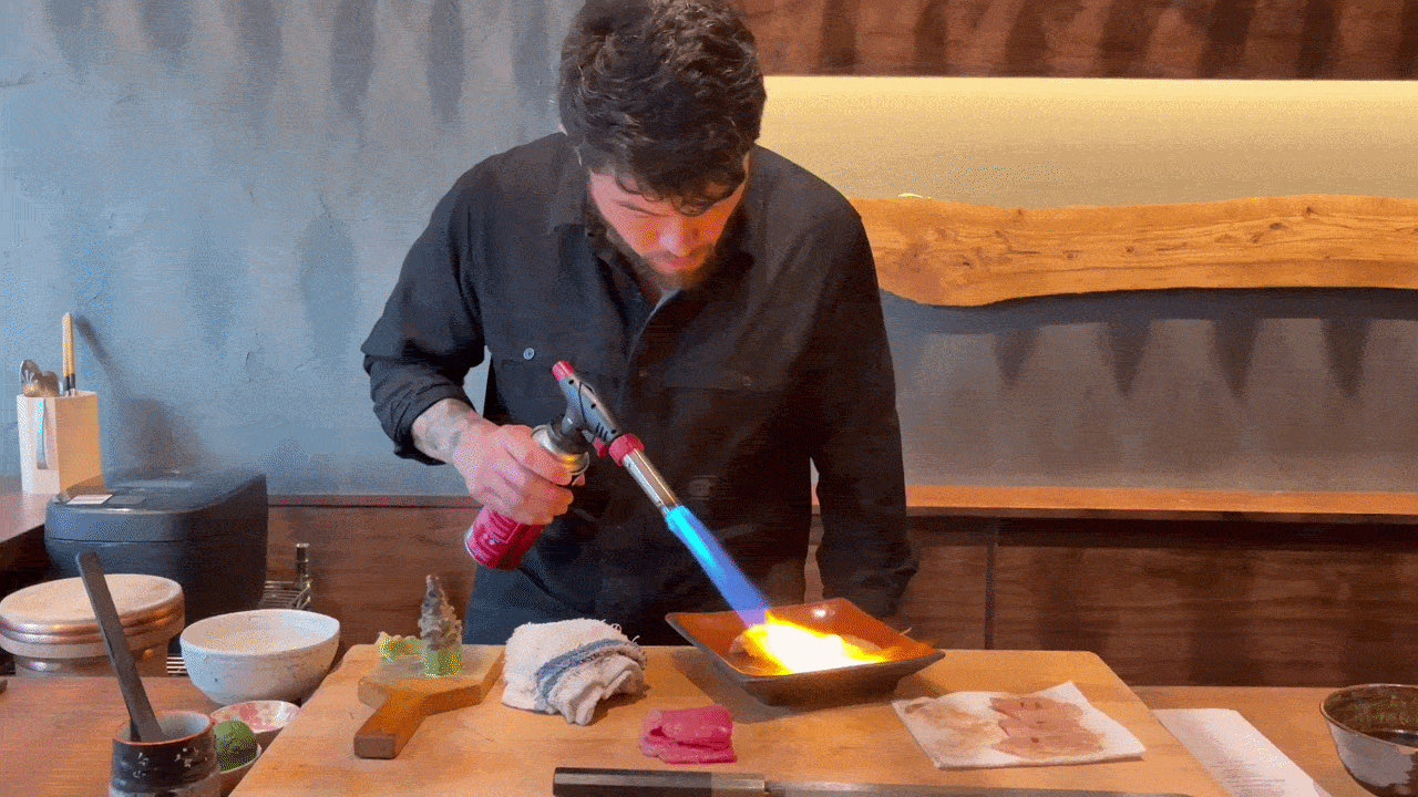 A man in a black shirt leans over a wooden counter, using a handheld blowtorch to heat an orange dish; a blue flame glows as tools, towels, and bowls lie nearby in a restaurant.