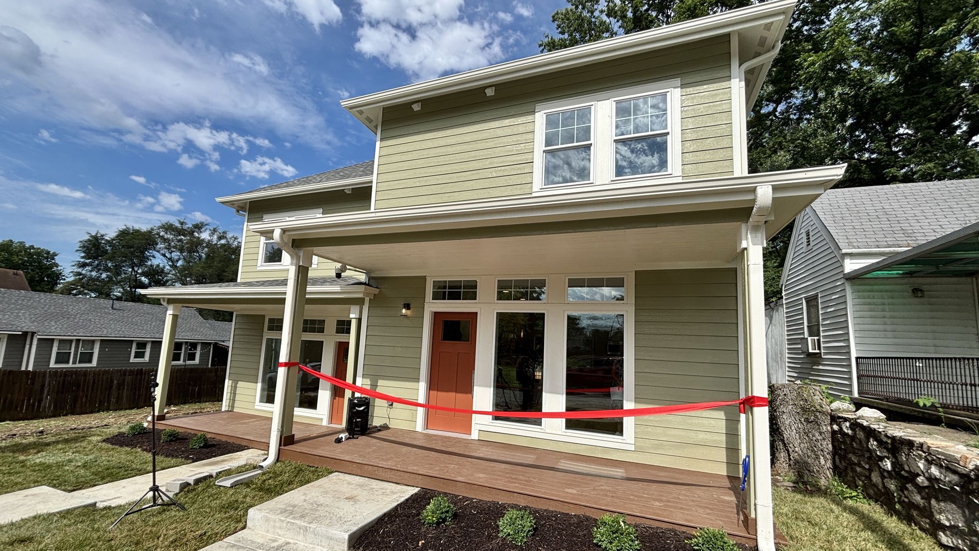Two-story house with sage-green siding and white trim, orange front door, and large front windows under a covered porch. Red tape blocks the entry as work continues; blue sky and trees.