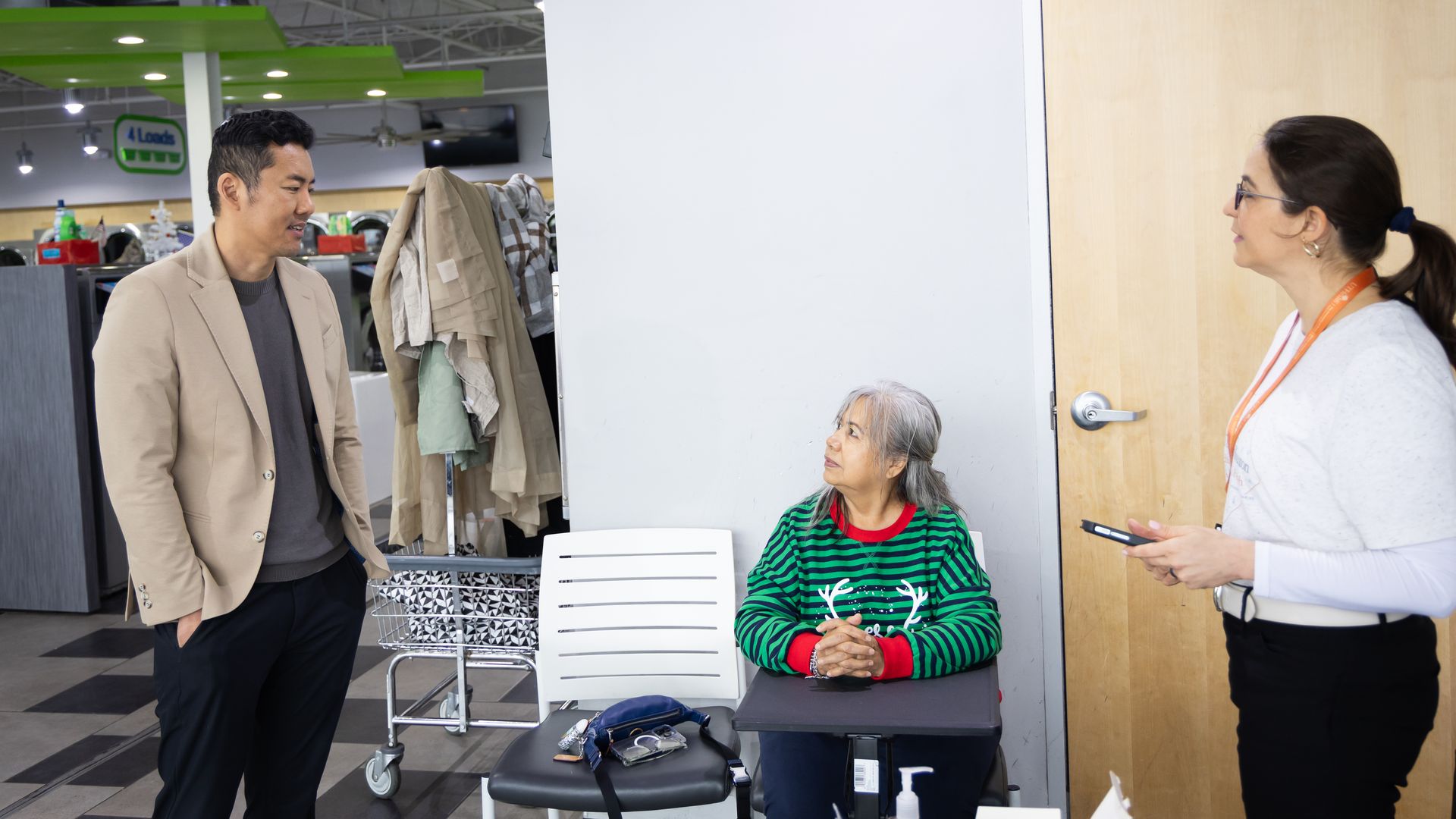 Inside a retail store, a man in a tan blazer talks to an employee wearing an orange lanyard while an elderly woman in a green striped sweater sits at a desk; a shopping cart and coats are nearby, with a Lowe's sign visible in the background.