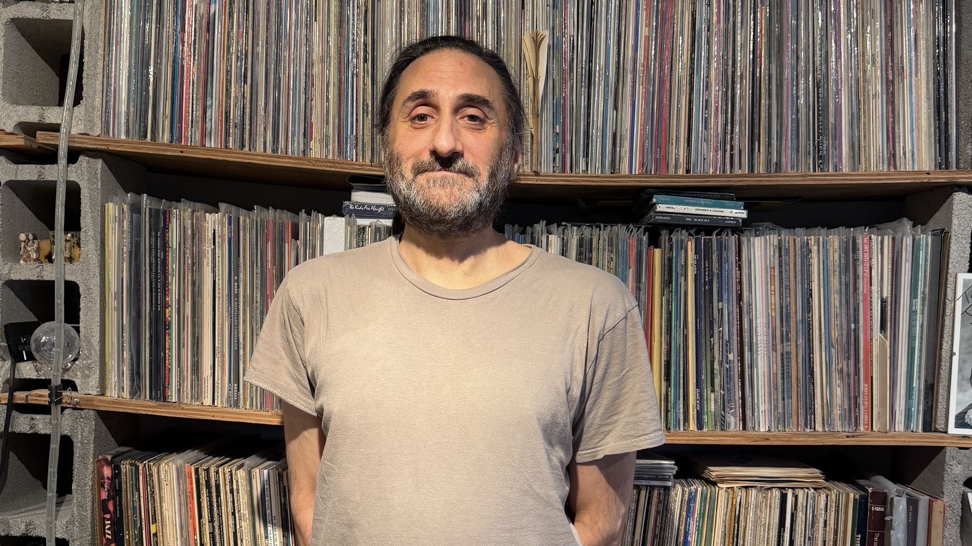 A bearded man with dark hair tied back stands before tall shelves filled with vinyl records; concrete blocks and wooden shelves frame rows of colorful album sleeves.