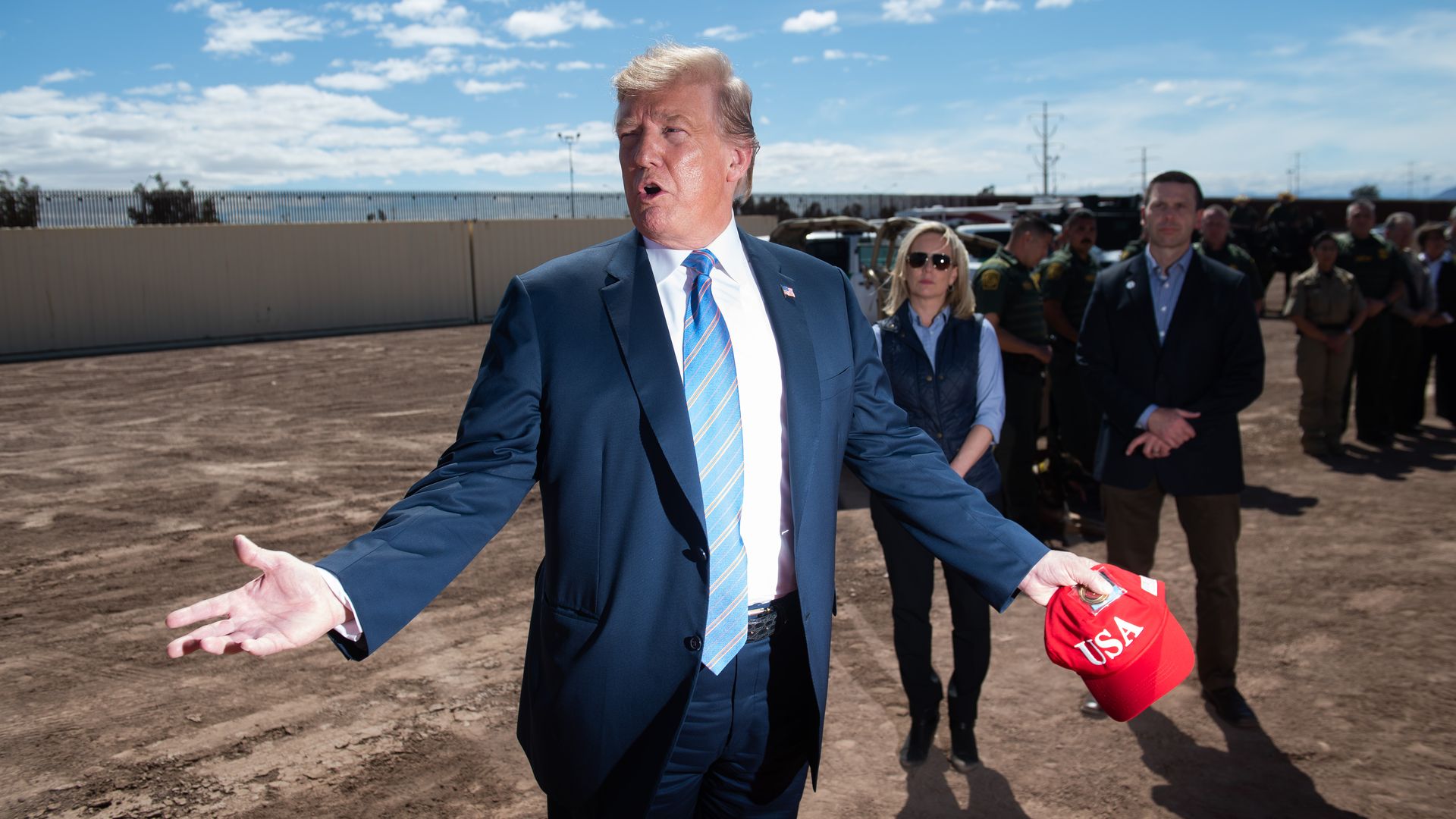 Donald Trump holding a red hat at the border, with former DHS Secretary Kirstjen Nielsen behind him