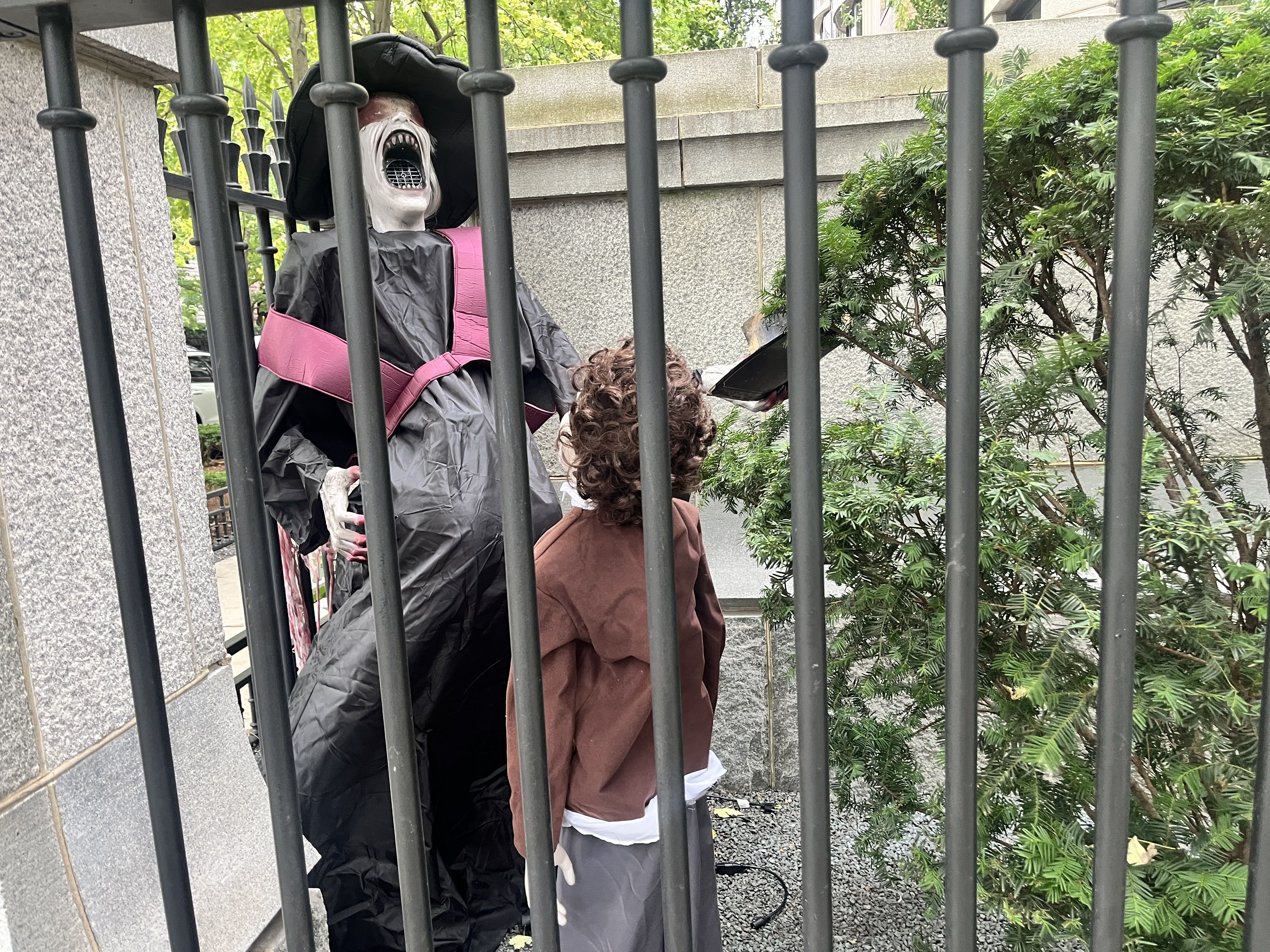 Life-size Halloween prop in a black robe and wide-brimmed hat, pale screaming face with an open mouth and teeth, pink sash across the chest, behind gray iron bars; stone wall and greenery in the background.