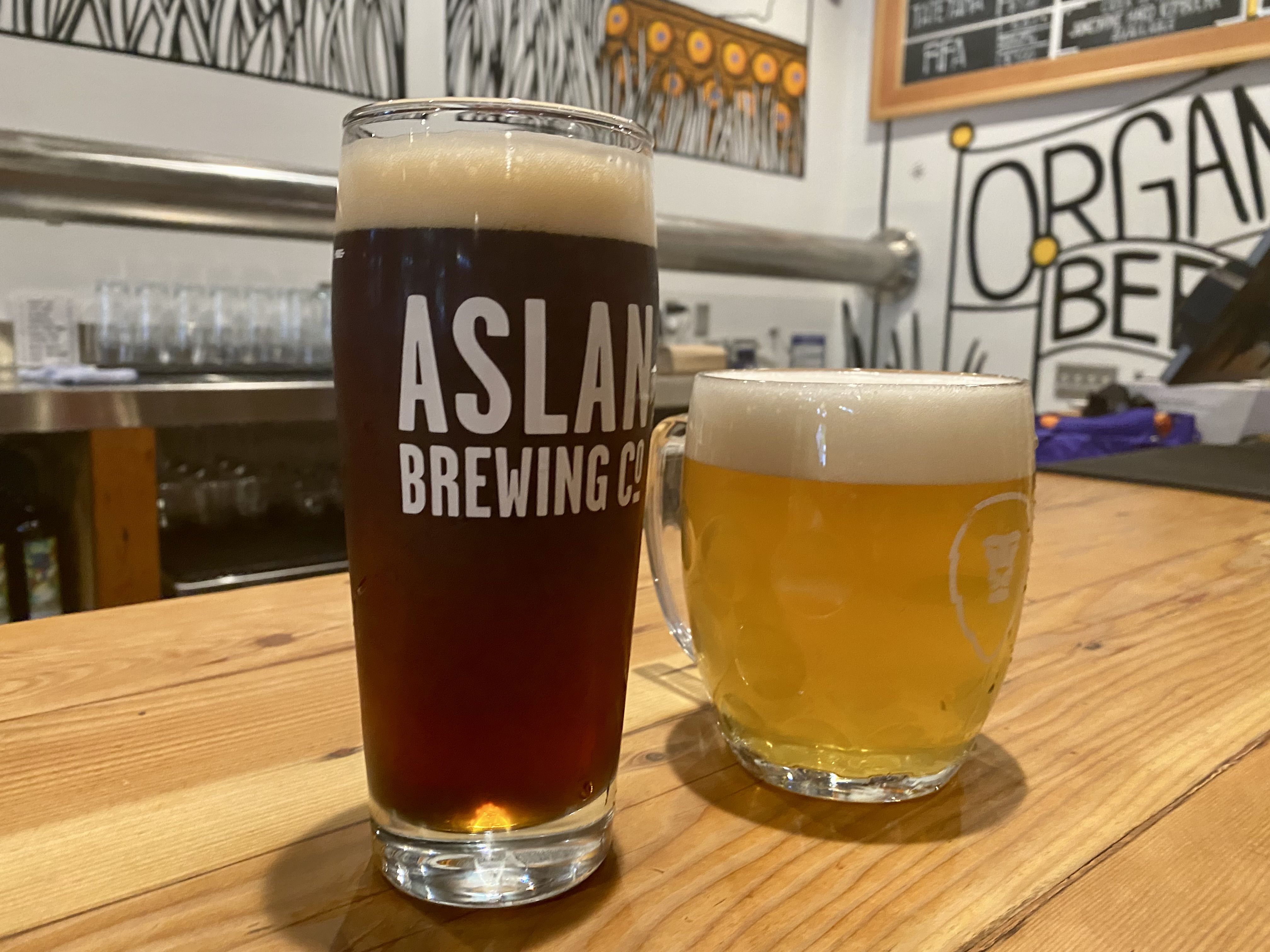 A brown beer in a tall glass and a lighter colored beer in a shorter glass, both labeled "Aslan Brewing," sitting on a wooden bar.