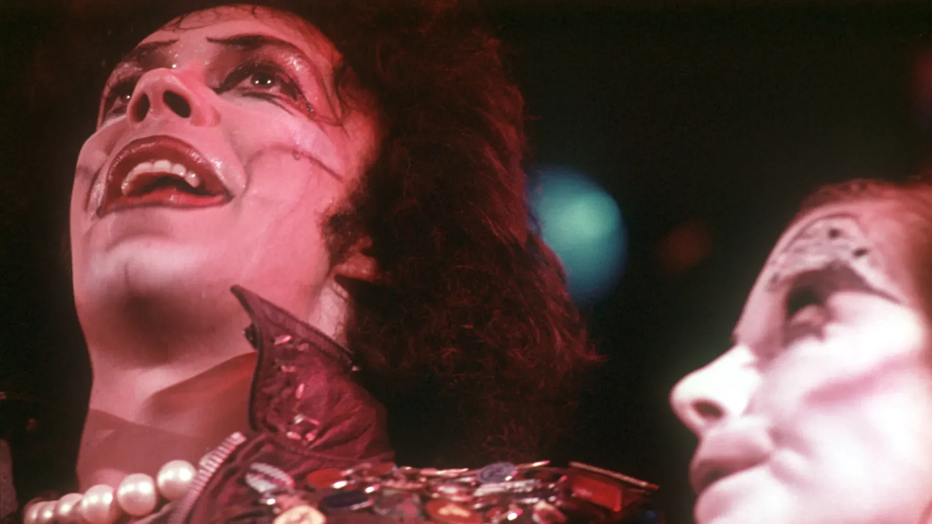 Close-up of two performers in dramatic stage makeup under red lighting: left figure with pale skin, dark eye makeup, open mouth and pearl necklace; right figure with white mask-like makeup.