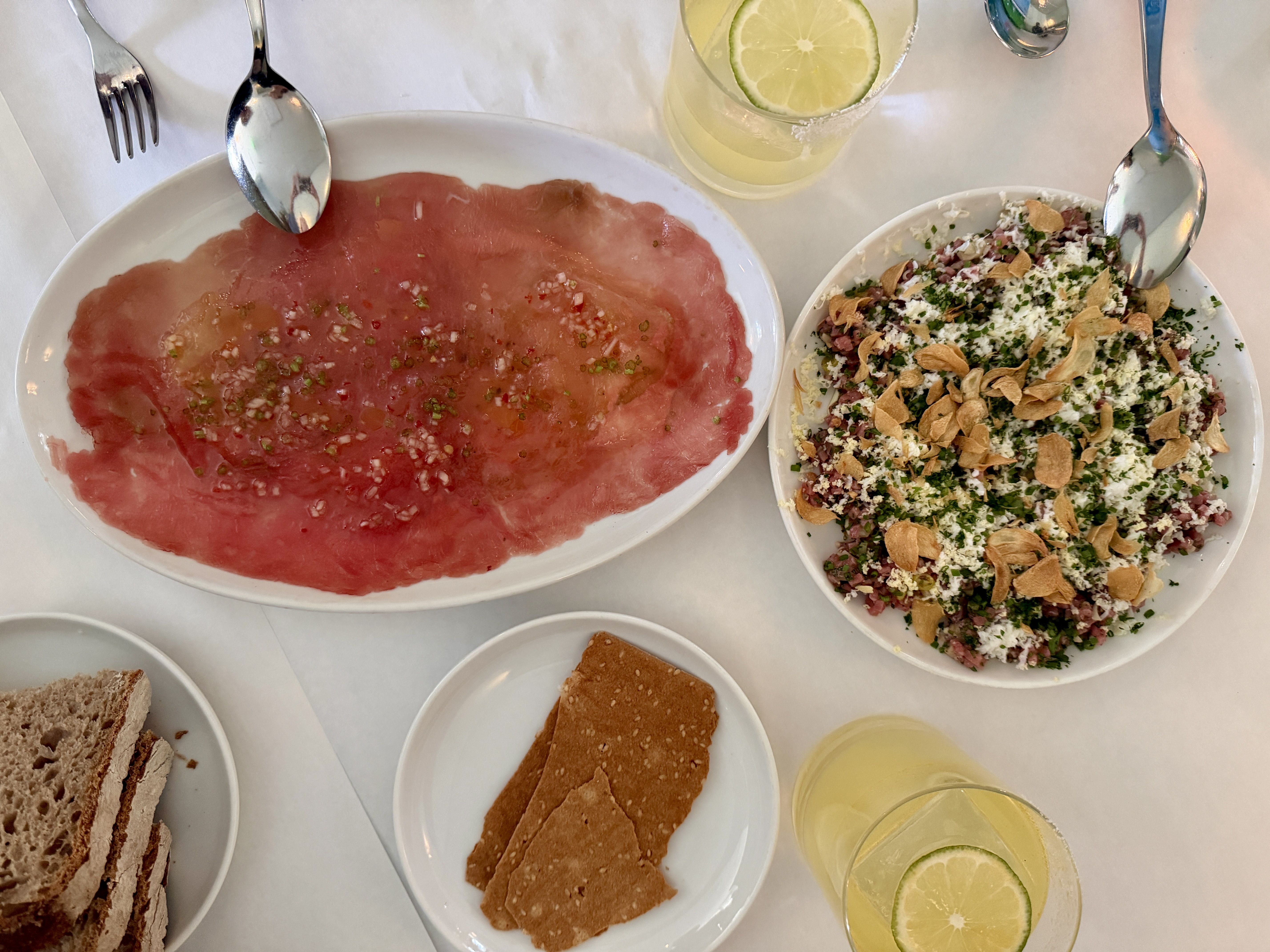 Top-down view of a table: white plate of beef carpaccio sprinkled with onions and herbs, another bowl of chopped salad with feta and almond slices, two lemon drinks, and nearby forks and spoons.
