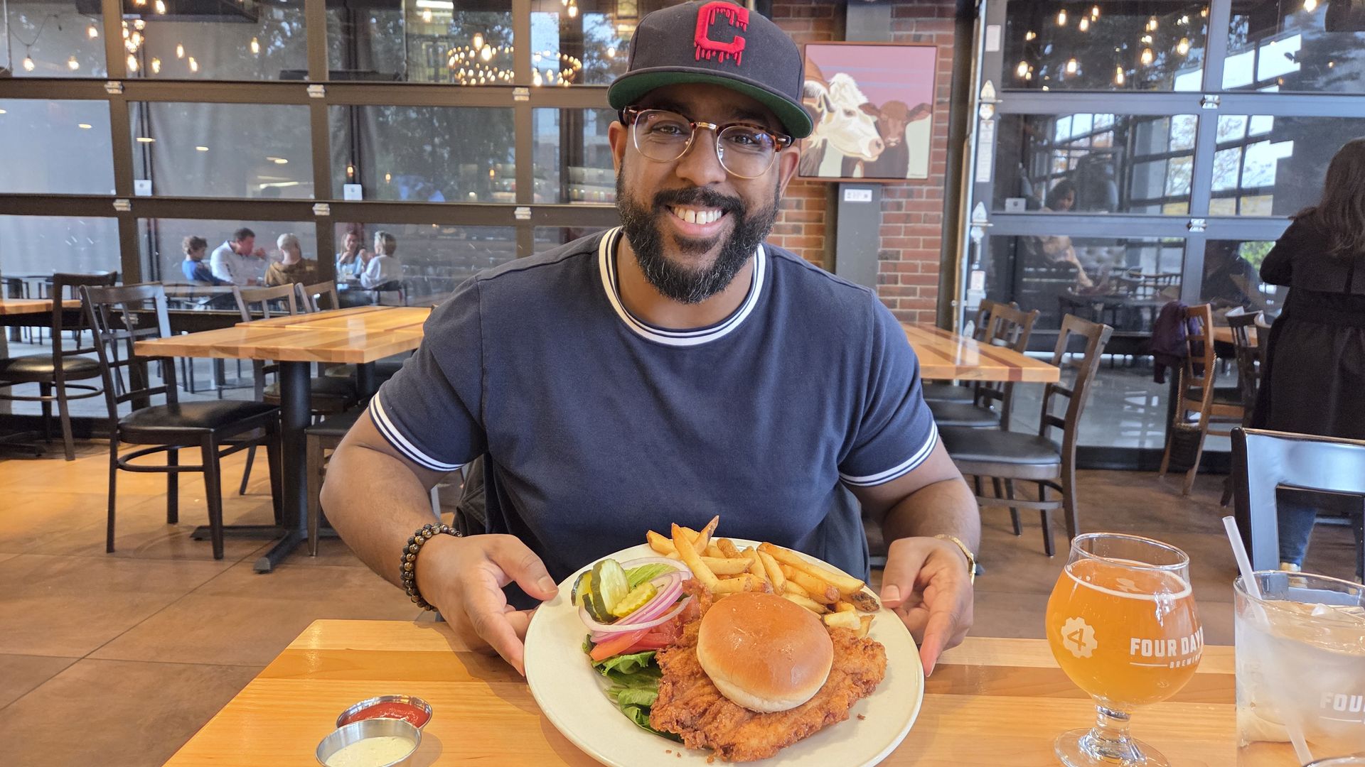 Smiling man wearing glasses and a black cap with red C sits at a wooden table in a restaurant, holding a plate with fried chicken sandwich, fries, and vegetables. Two drinks are on the table.