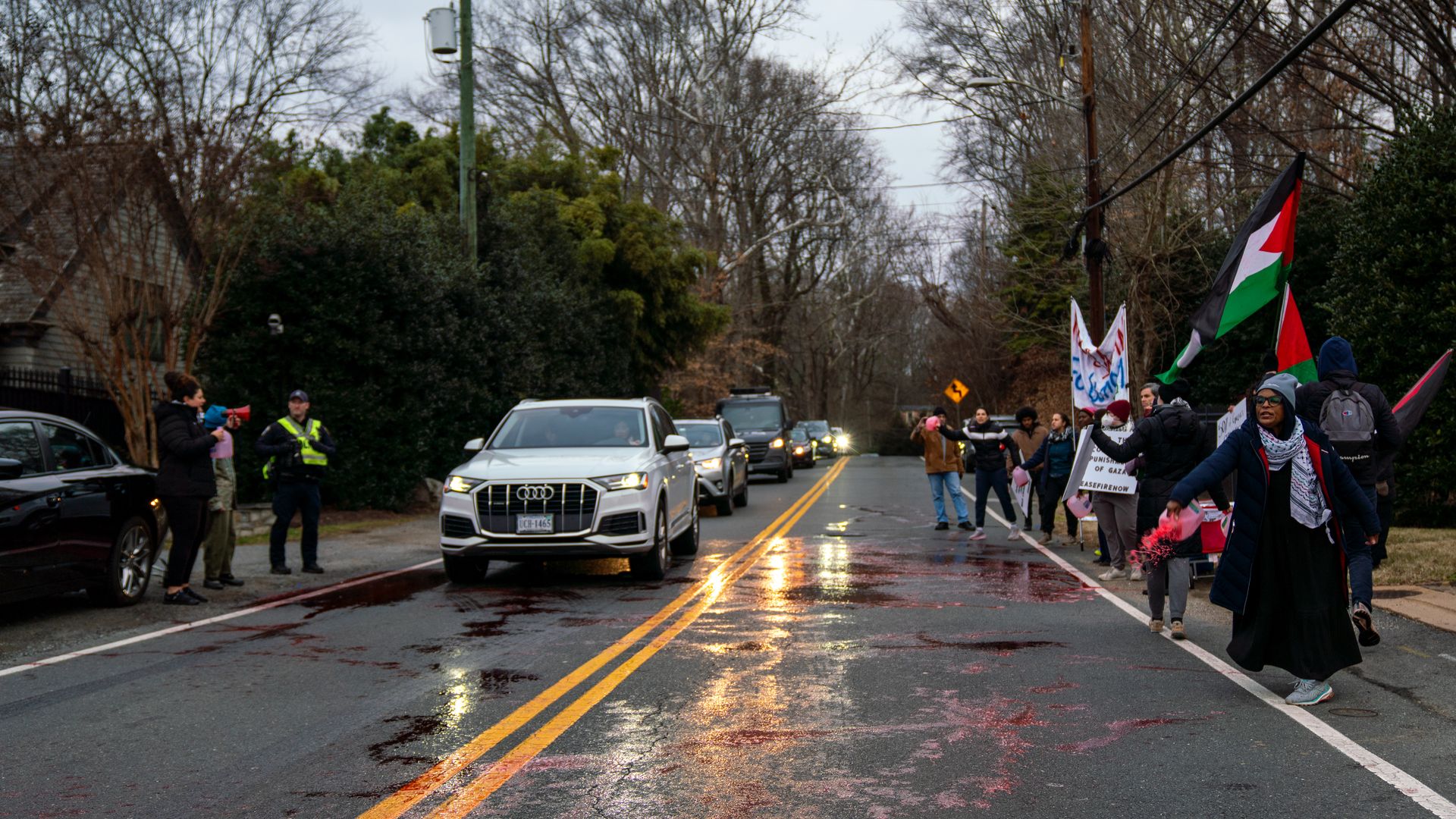 Protesters waving Palestine flags stop traffic while fake blood stands in the street.