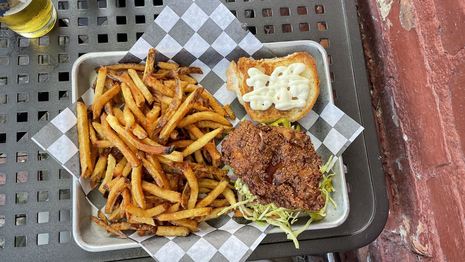 Top-down view of a metal tray with crispy fries and a fried chicken sandwich on a toasted bun topped with mayo; shredded lettuce underneath and a beer glass at the top left.
