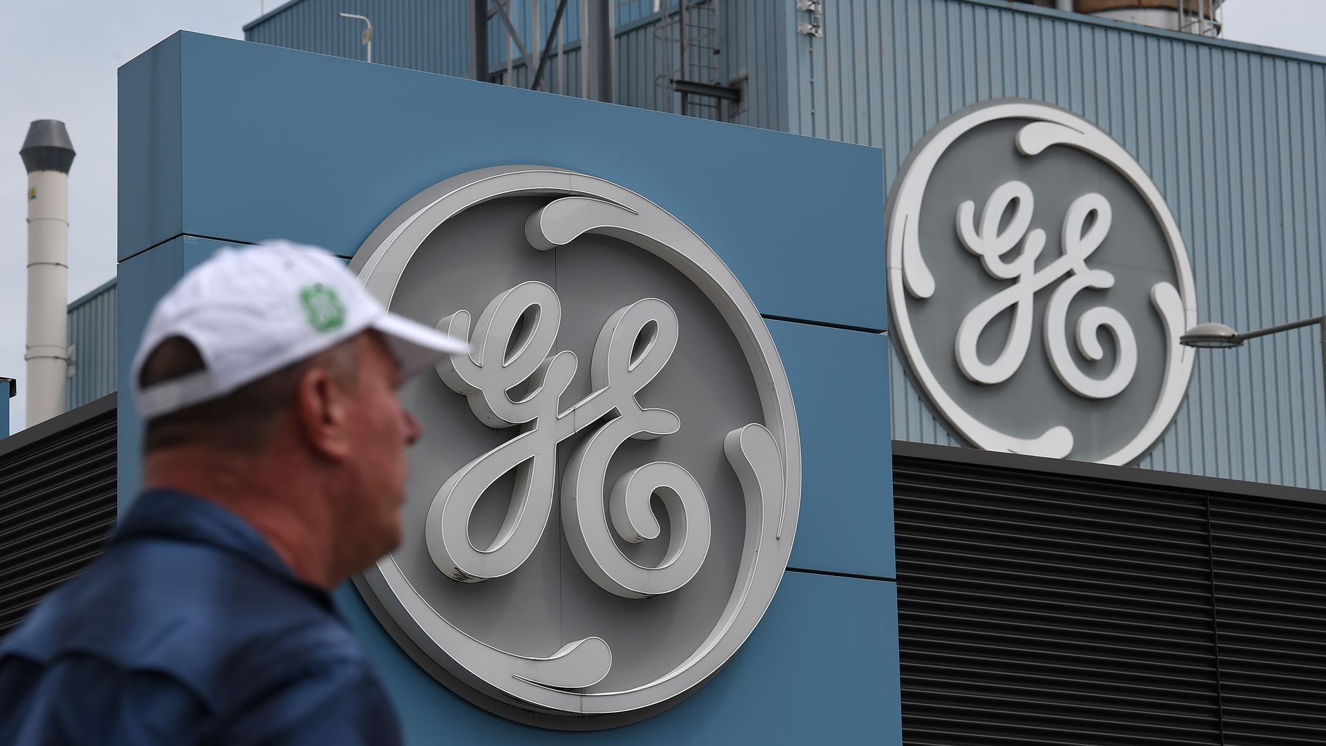 In this image, a man wearing a baseball cap walks in front of a building with a giant General Electric logo.