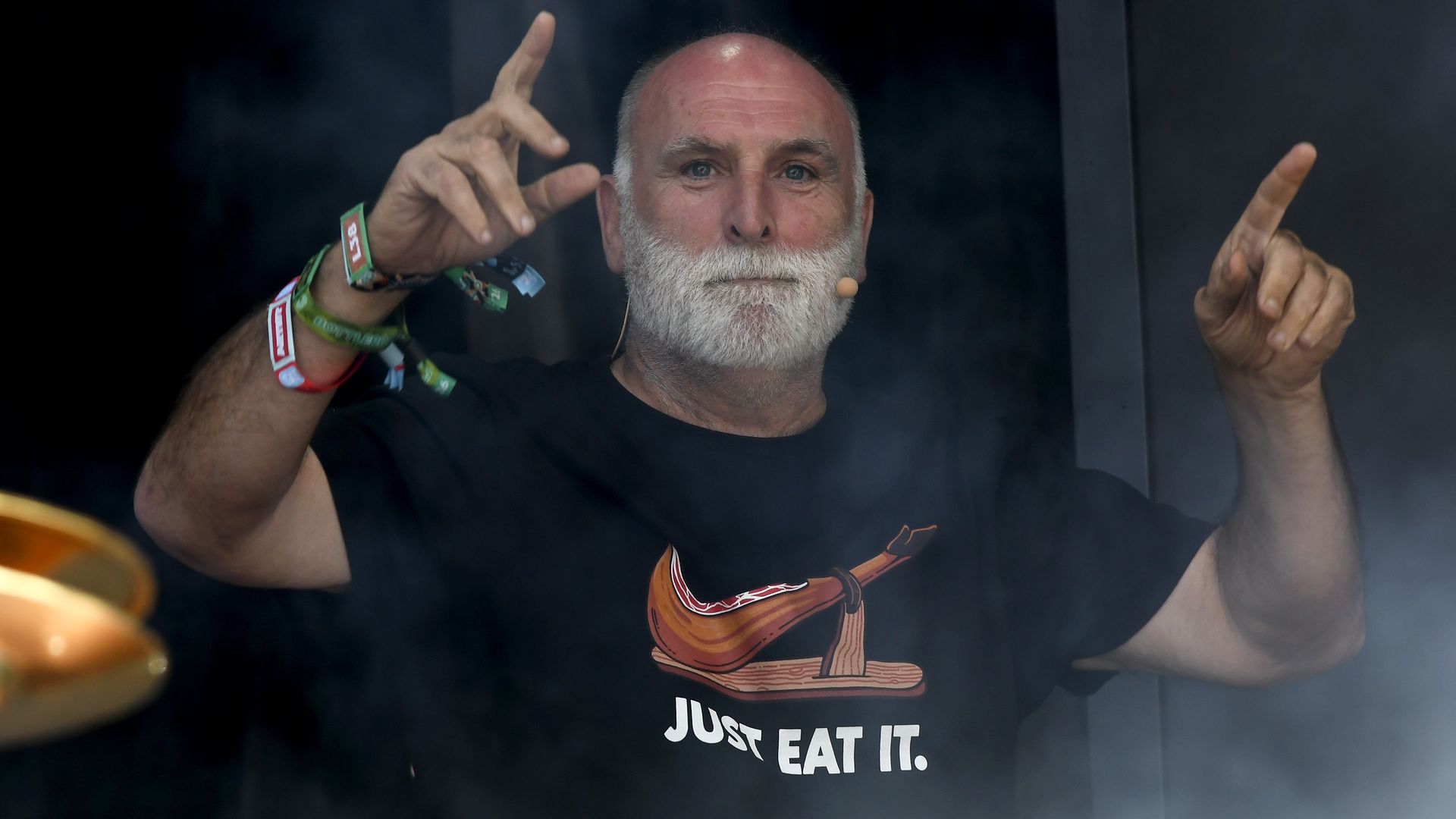 Close-up of a bearded, bald man in a black shirt that reads "JUST EAT IT," wearing a headset mic. He raises both hands with extended fingers; smoky dark background and a bowl in the corner.