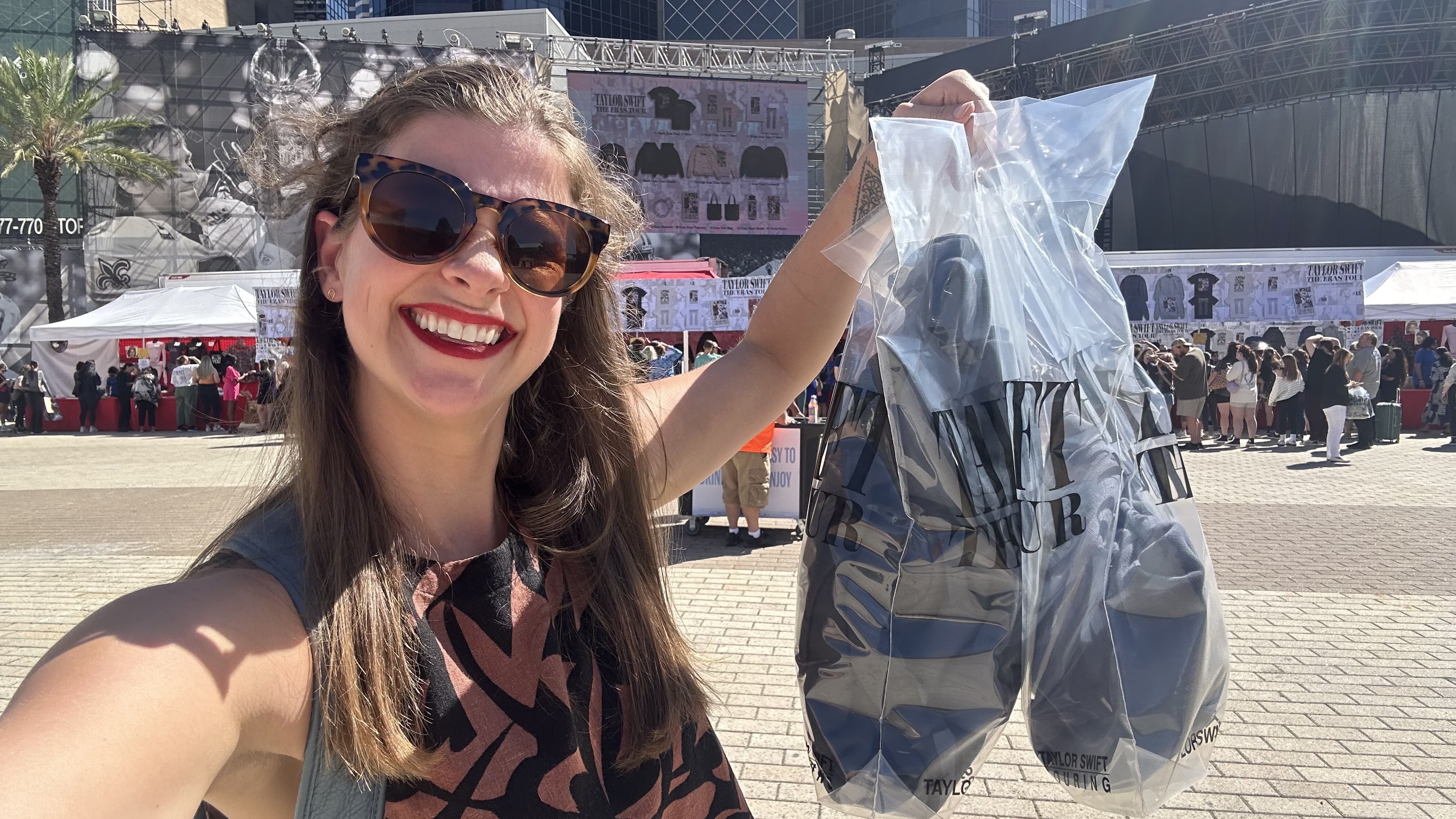 A selfie of a woman as she holds up two shopping bags. Behind her is the Taylor Swift merchandise area.